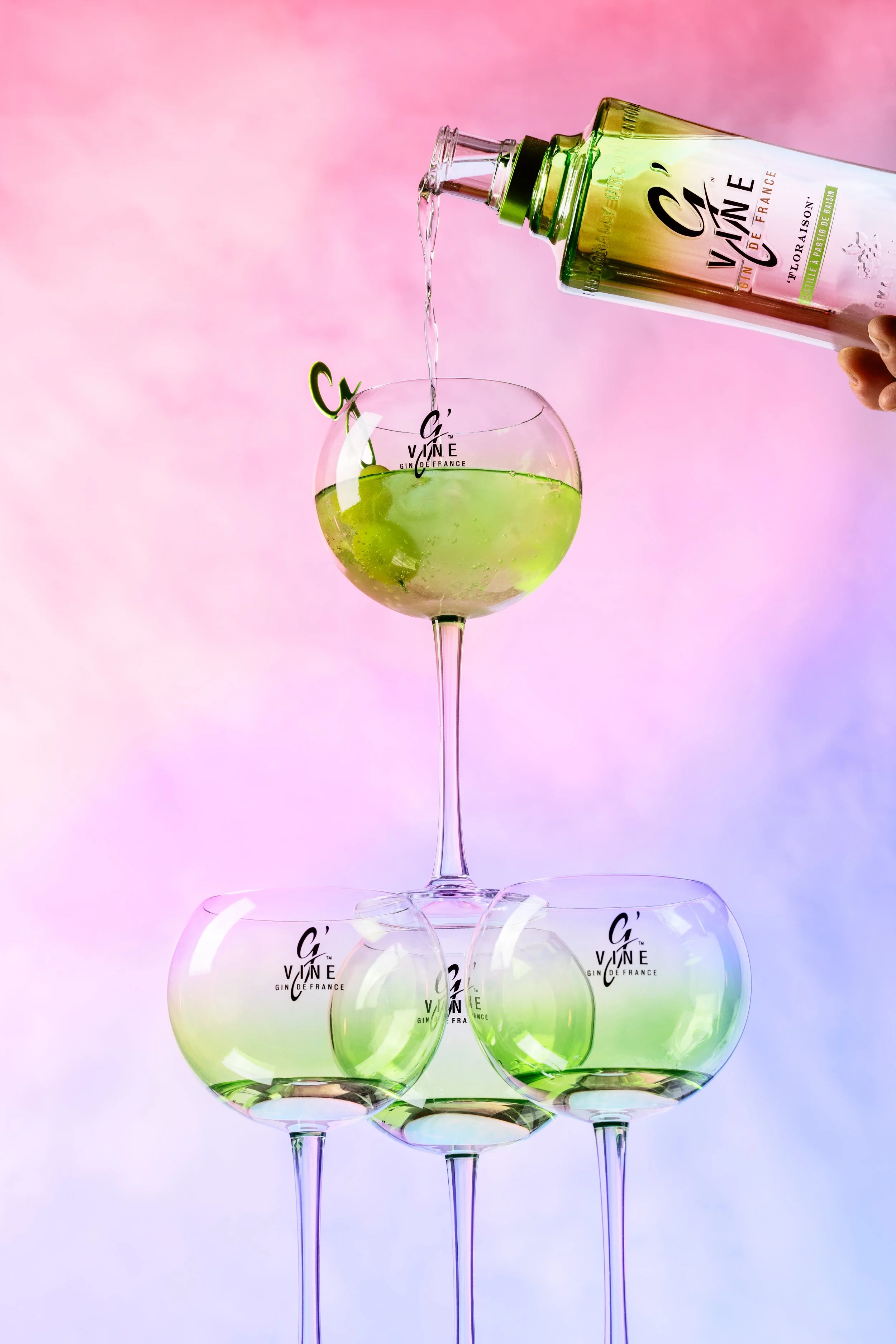 Verre de cocktail vert avec tranche de citron, verre de vin vert en dessous, bouteilles de gin avec étiquettes « G'Vine » en arrière-plan, sur un fond pastel dégradé.