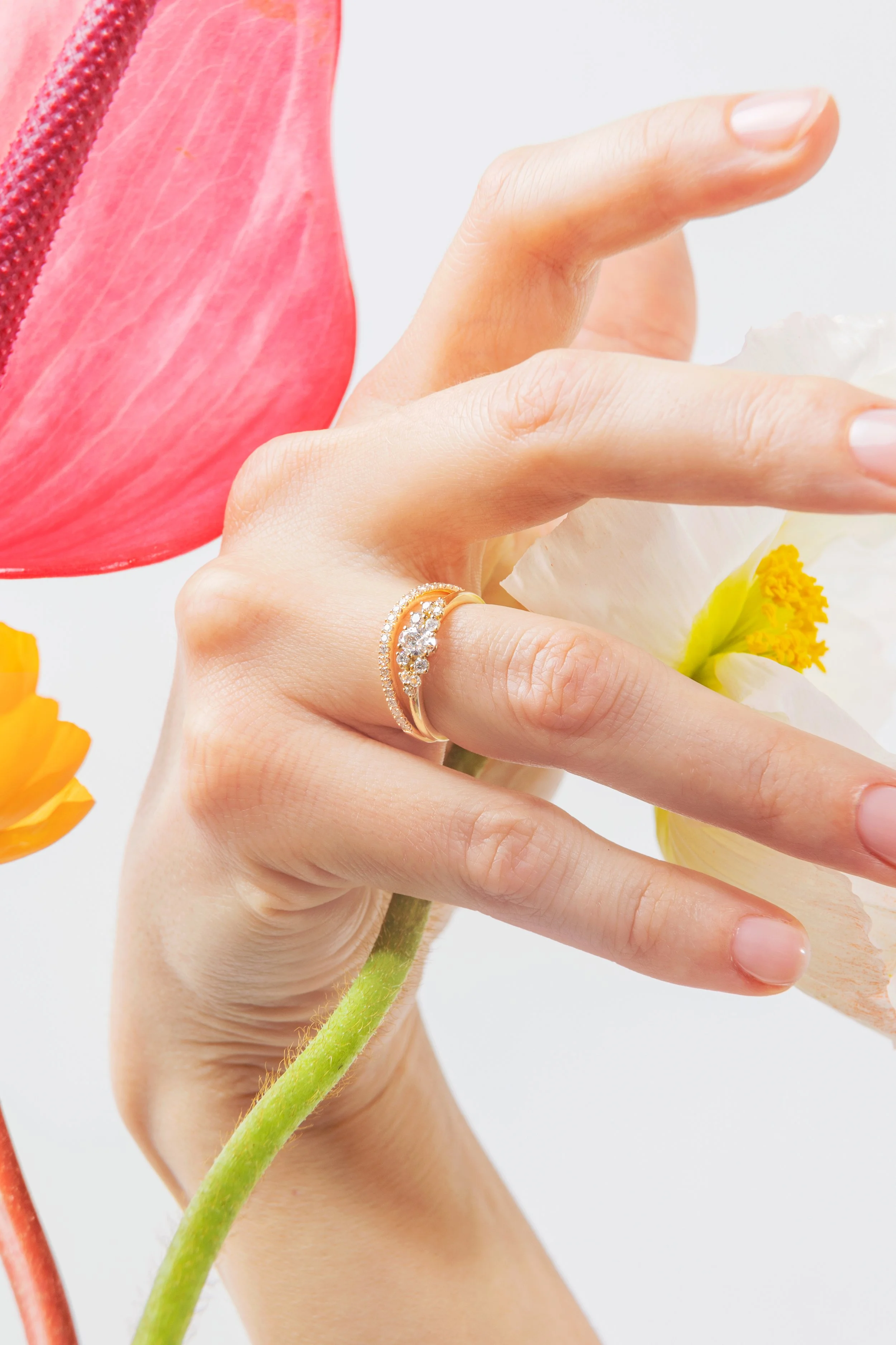 Une main portant une bague en or avec des pierres précieuses, tenant une fleur blanche avec un centre jaune. D'autres fleurs colorées sont visibles en arrière-plan.