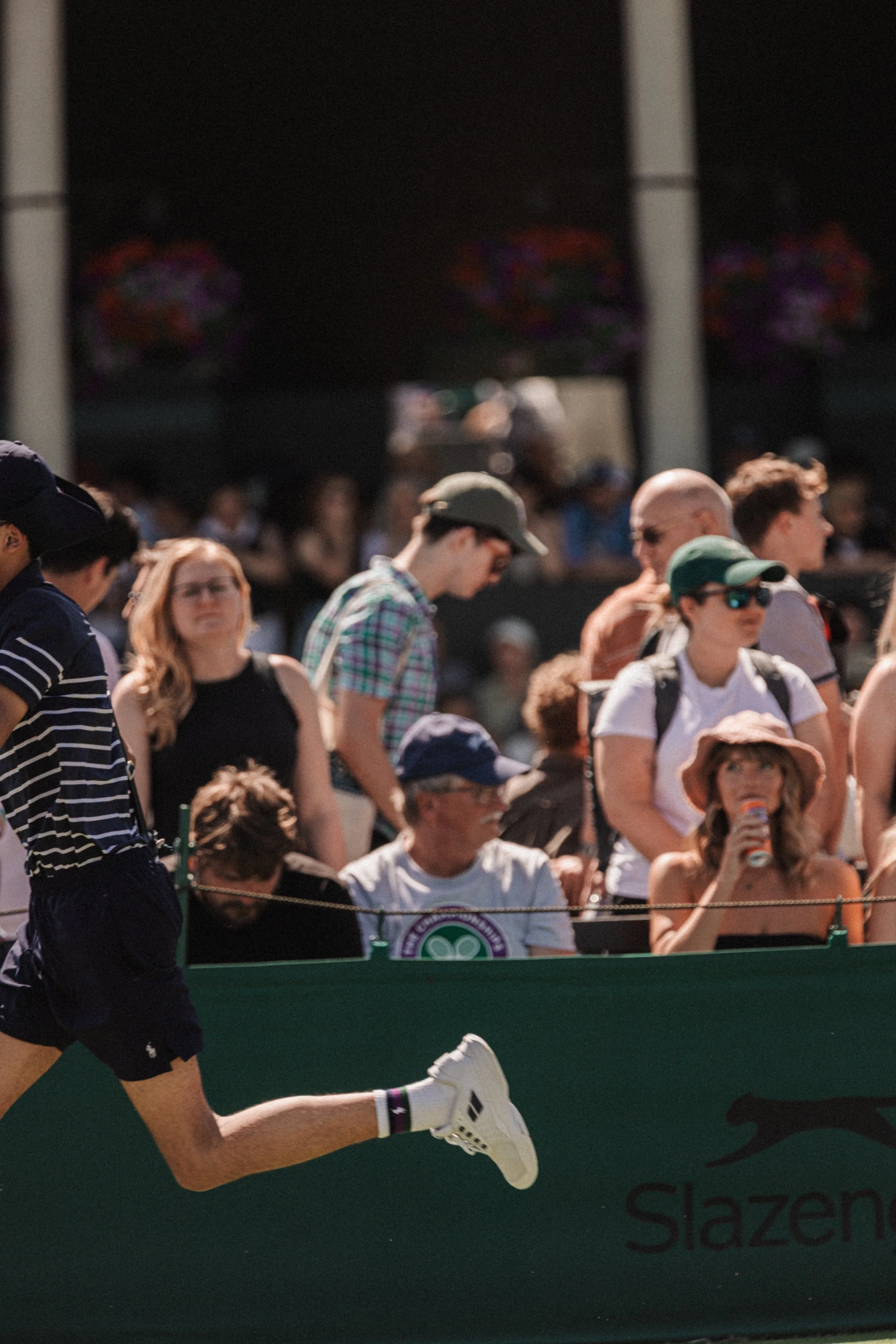 Vue d'ensemble de spectateurs d'un match de tennis lors d'un tournoi, avec plusieurs personnes debout dans une zone extérieure, certaines portant des chapeaux et des lunettes de soleil, derrière une barrière verte portant le logo et une inscription p
