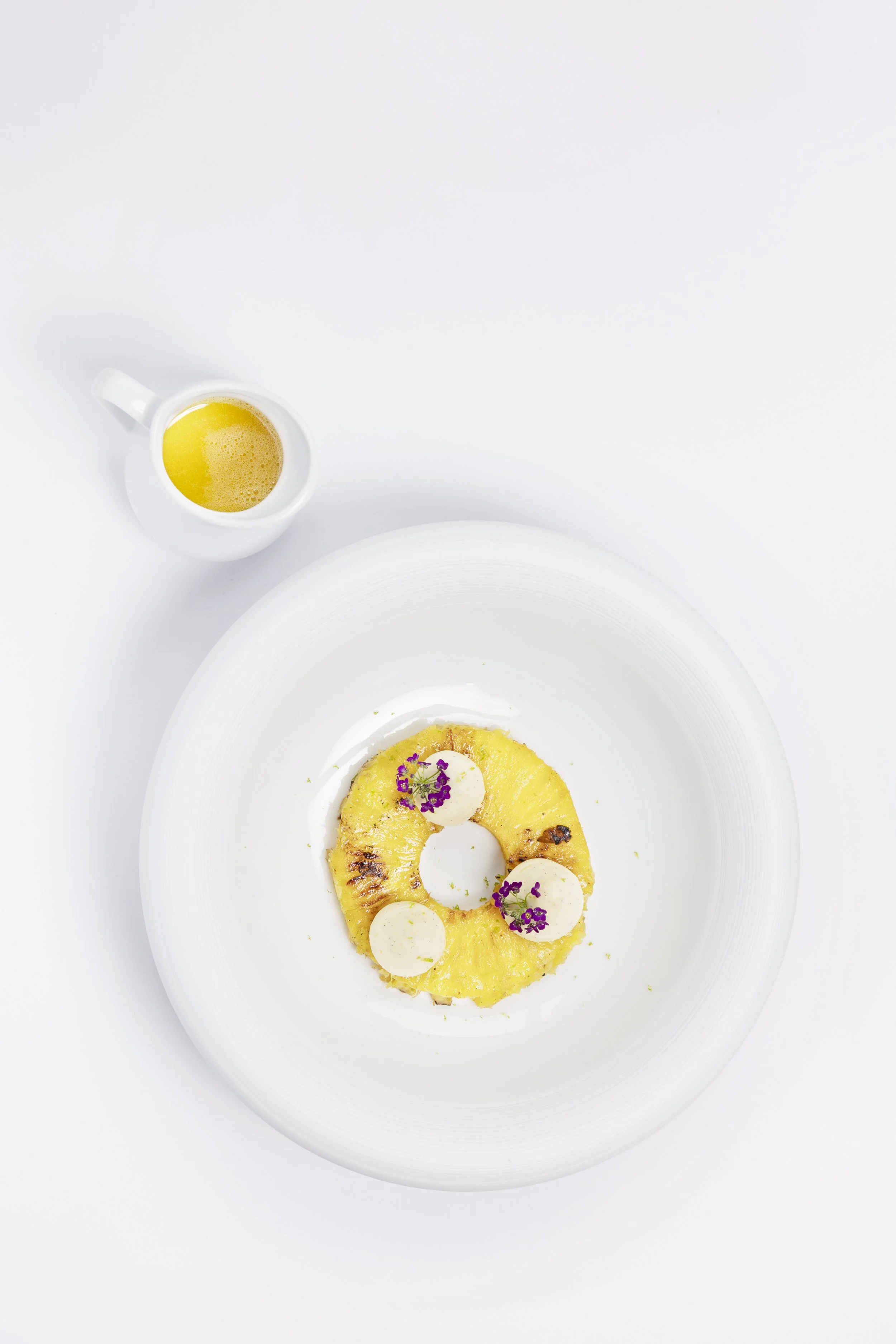 Ananas grillée avec des tranches de fromage et des fleurs pour la décoration, servie sur une assiette blanche, accompagnée d'une tasse de jus de fruit jaune, sur fond blanc minimaliste.