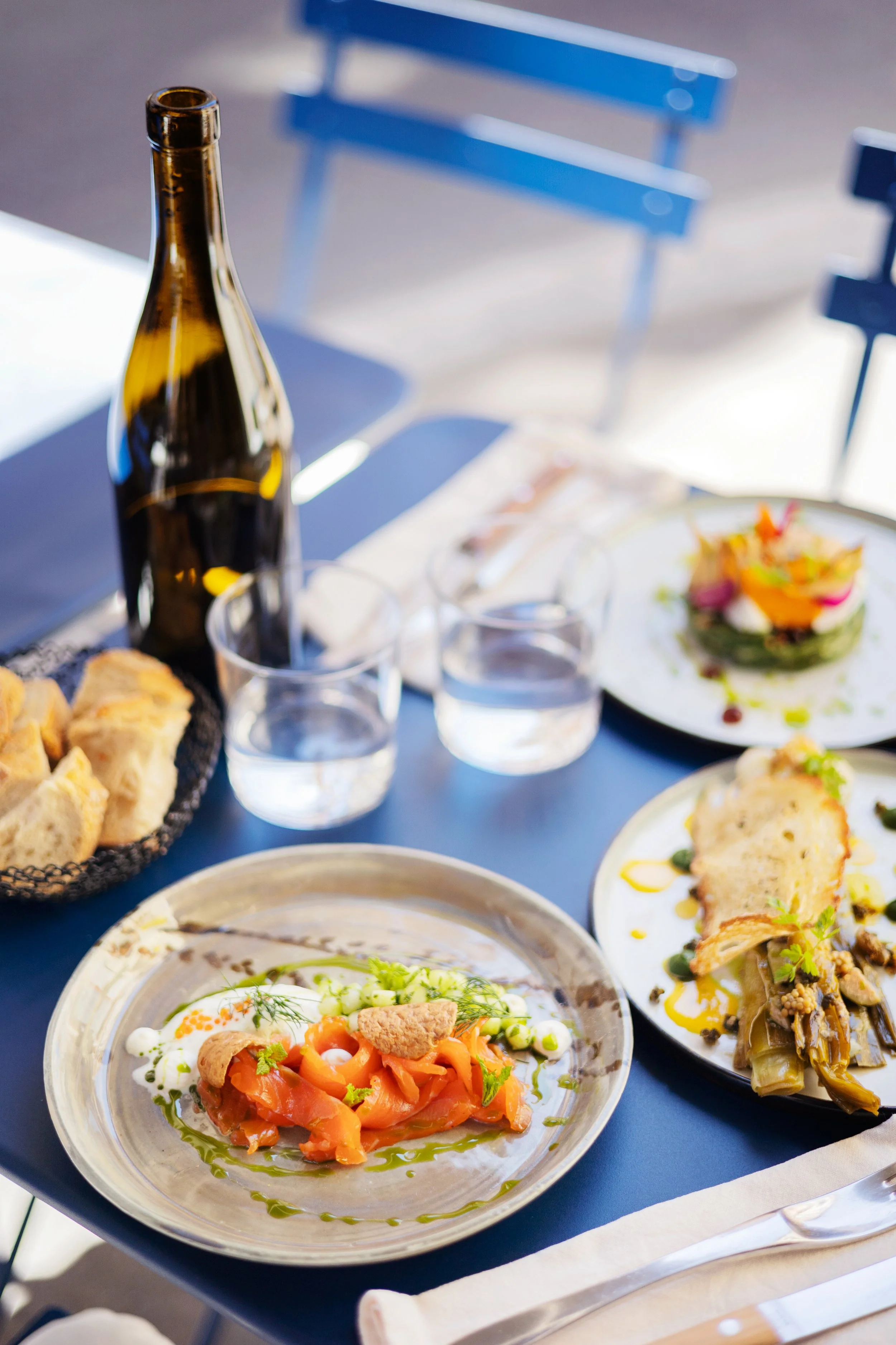 Assiette de carpaccio de saumon avec des herbes fraîches, assiette de gratin, présentation de plats variés sur une table bleue avec des verres d'eau.
