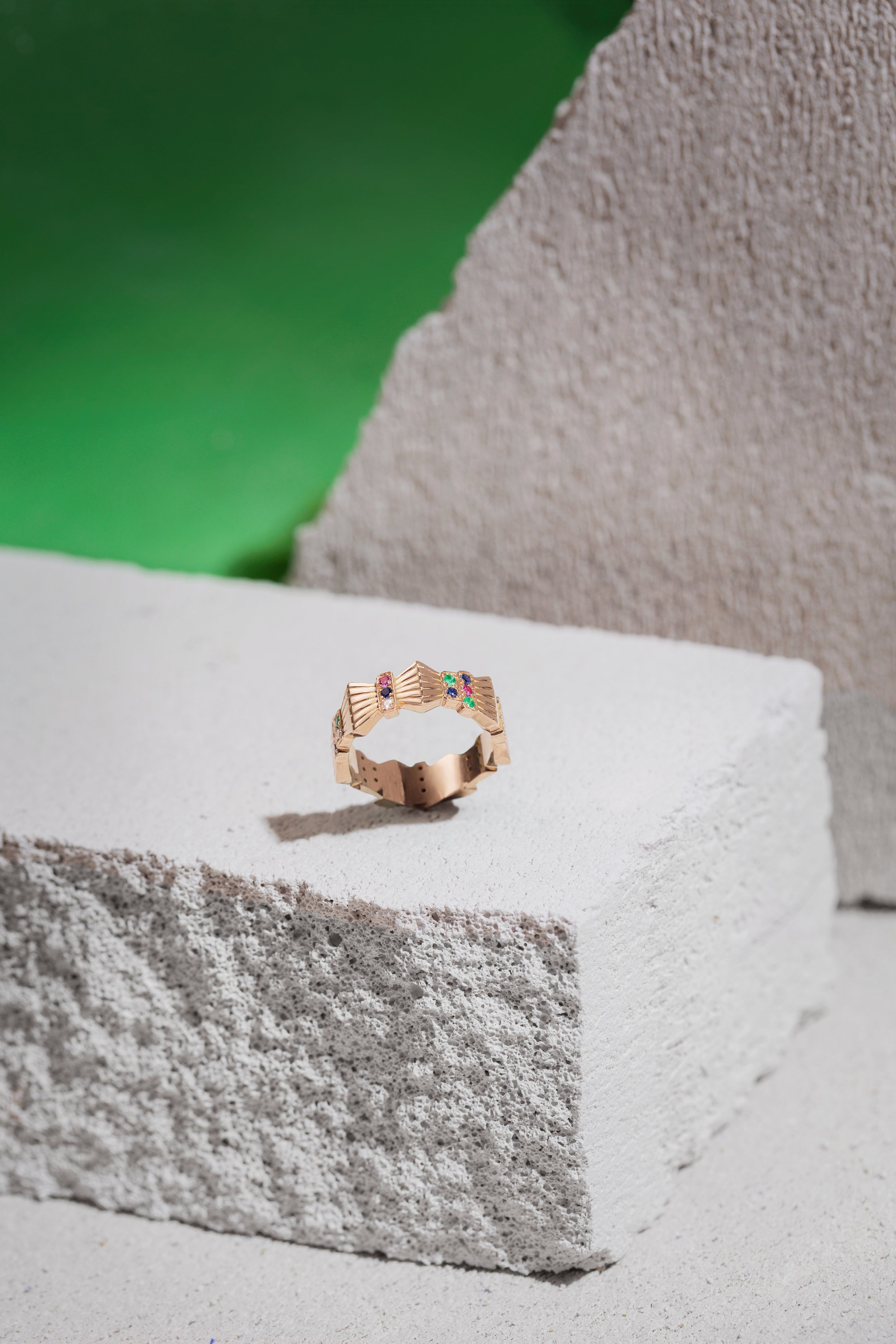 une bague en or rose avec des pierres précieuses colorées posée sur un bloc de pierre brute, avec un fond en dégradé de vert, beige et blanc