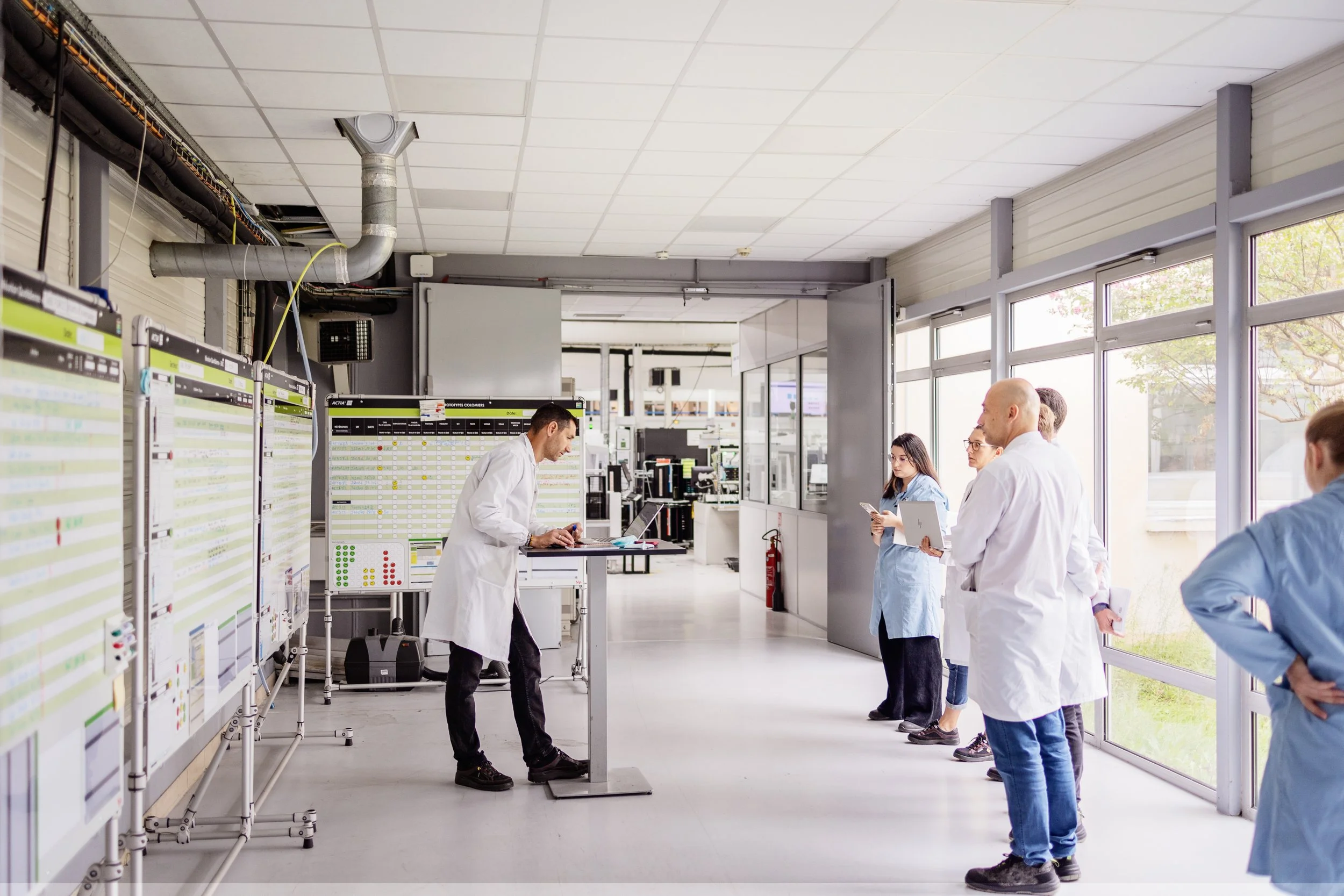 Des professionnels de la santé en blouses blanches à l'intérieur d'une salle de laboratoire ou d'hôpital, certains utilisant des ordinateurs ou des tableaux, avec de grandes fenêtres laissant entrer la lumière naturelle.