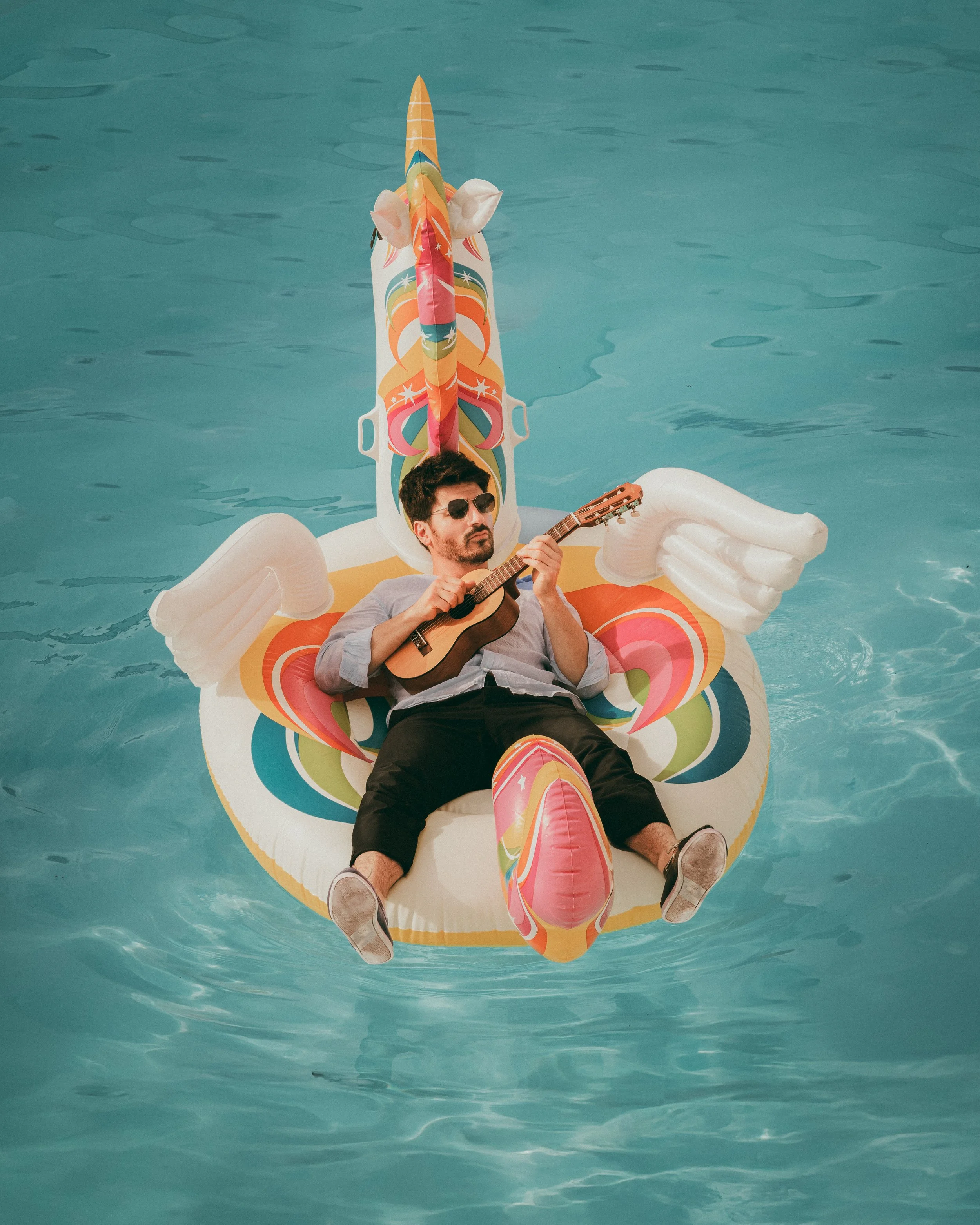 Une personne joue de la guitare tout en étant allongée sur une bouée en forme de licorne dans une piscine.