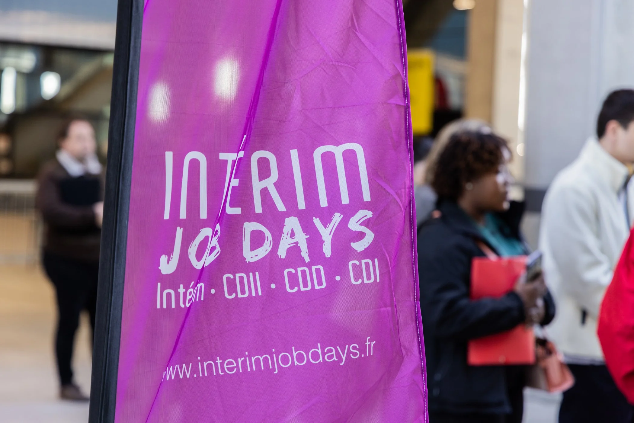 Une bannière rose avec du texte blanc annonçant « INTERIM JOB DAYS » et un site web www.intermjobjdays.fr. En arrière-plan, des personnes attendent, certaines tenant des documents ou des cafés, dans un lieu probablement urbain ou une salle d'attente.