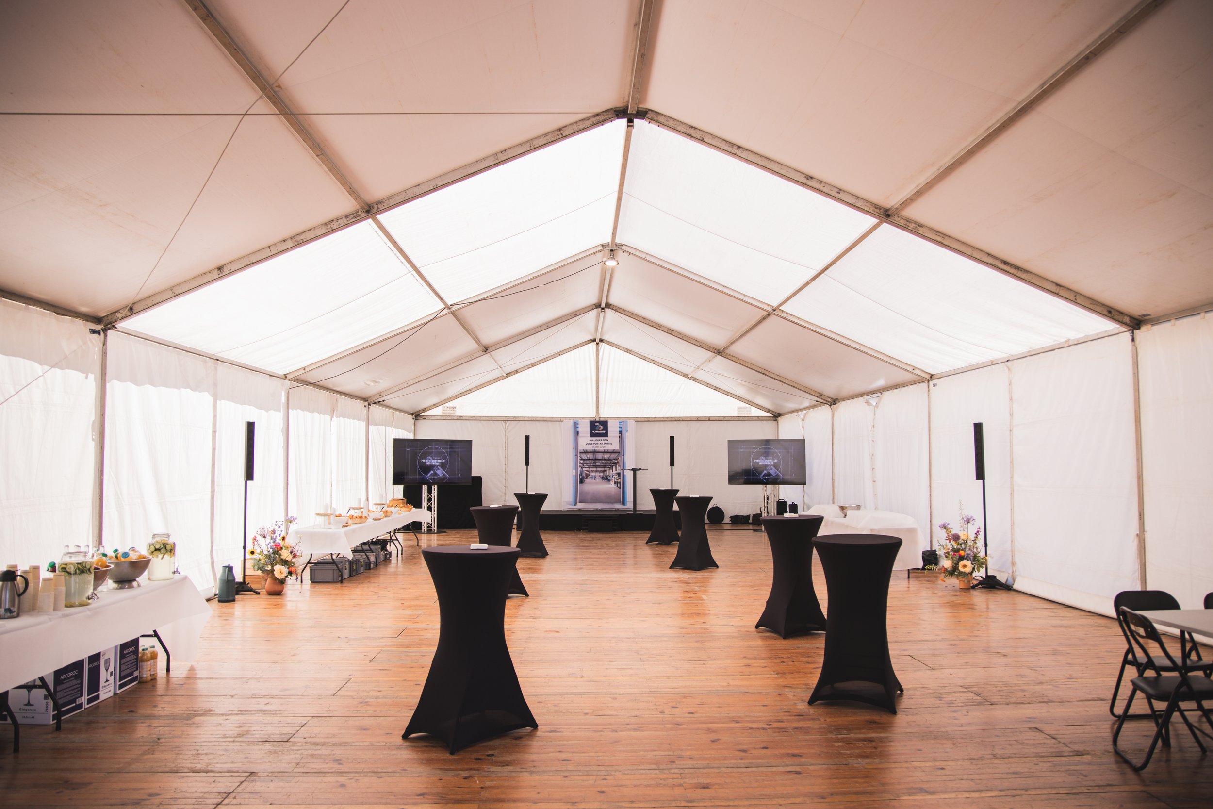Intérieur d'une grande tente blanche aménagée pour un événement, avec des tables hautes couvertes de décorations noires, des écrans, et diverses tables avec buffet et fleurs.