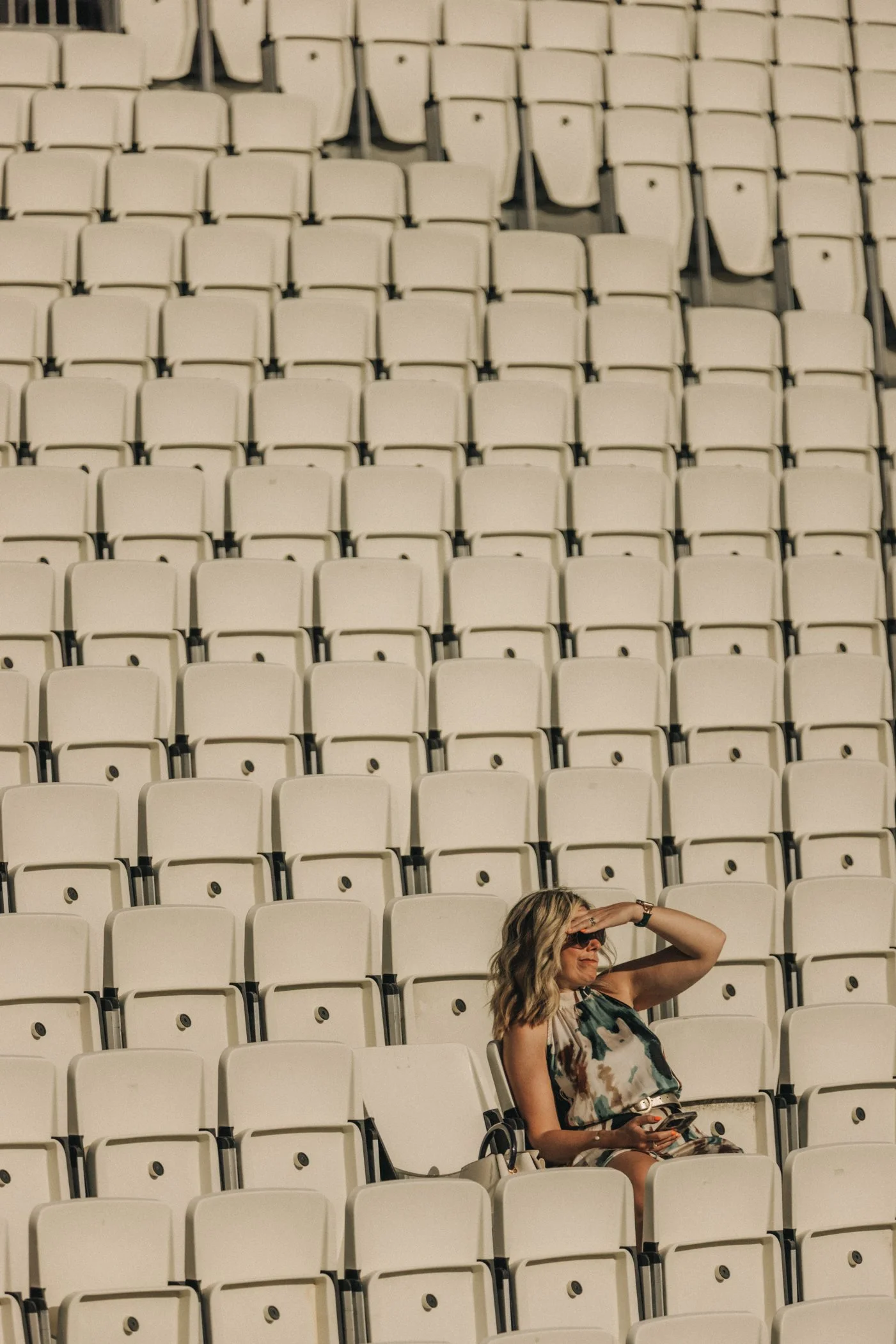 Une femme assise dans les sièges d'un stade vide, portant des lunettes de soleil et tenant un téléphone, avec un bras levé pour se protéger du soleil.