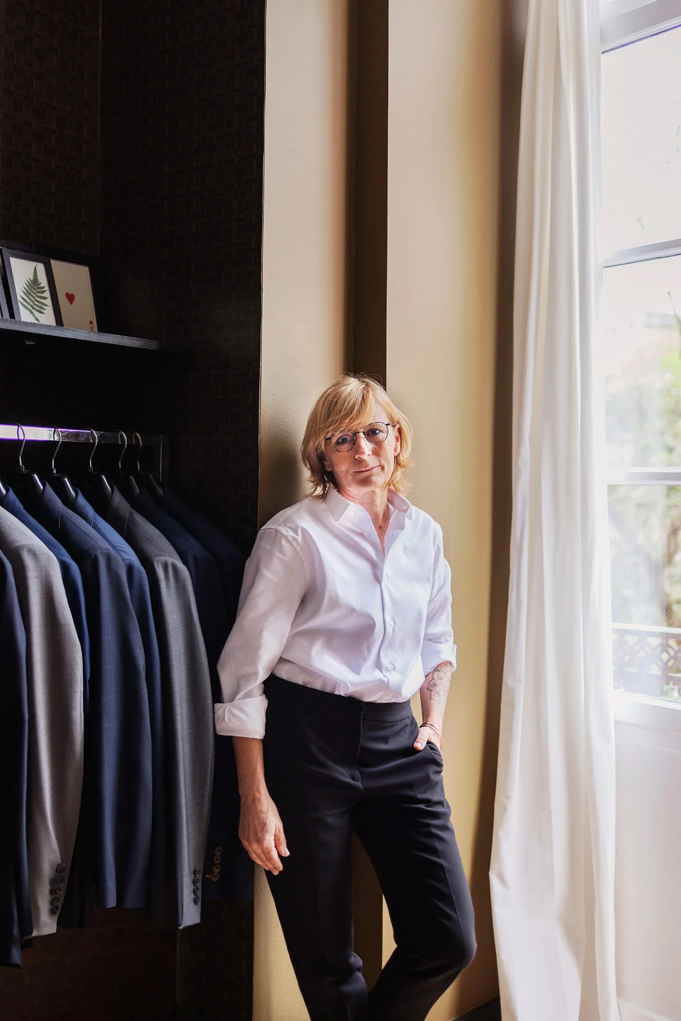 Femme blonde portant des lunettes, habillée d'une chemise blanche et d'un pantalon noir, posant dans une pièce lumineuse à côté d'une fenêtre avec des rideaux blancs, des vestes suspendues à côté.
