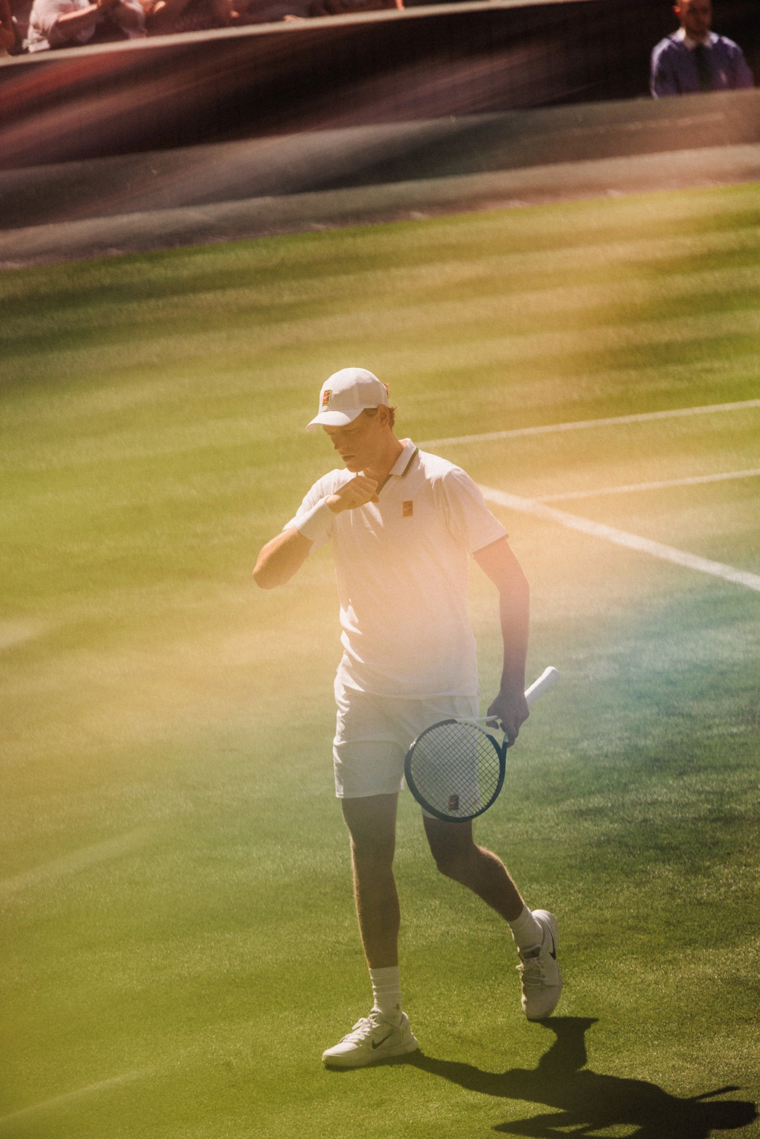Un jeune homme habillé en tenue de tennis, portant une casquette blanche, tenez une raquette de tennis dans une main, il marche sur un terrain de tennis en herbe.