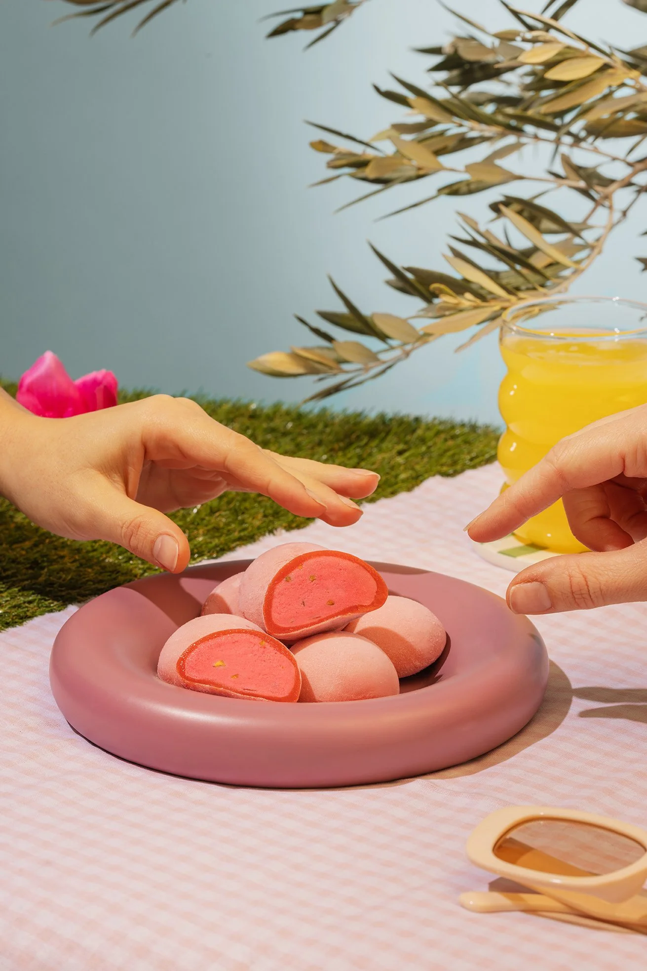 Gâteau en forme de rond, rose, avec un intérieur rose vif, présenté sur une assiette rose, avec deux mains qui tiennent une tranche. En arrière-plan, une bouteille de jus jaune, des branches d'olivier, une nappe rose et un fond bleu clair.