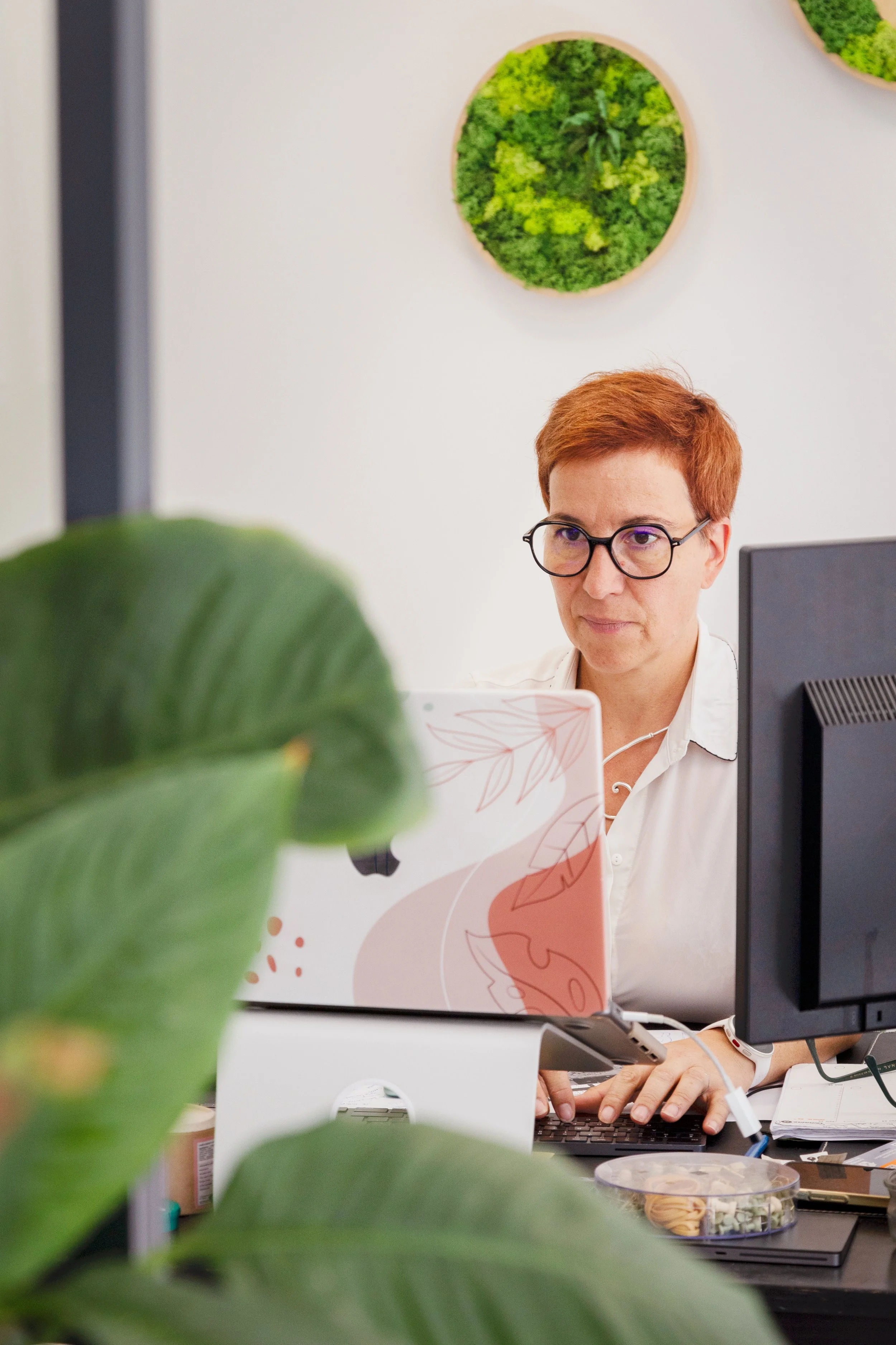 Une femme avec des lunettes rouges travaille sur un ordinateur dans un bureau avec un mur blanc, décoré de cadres avec des plantes vertes murales. Il y a une plante verte en premier plan.