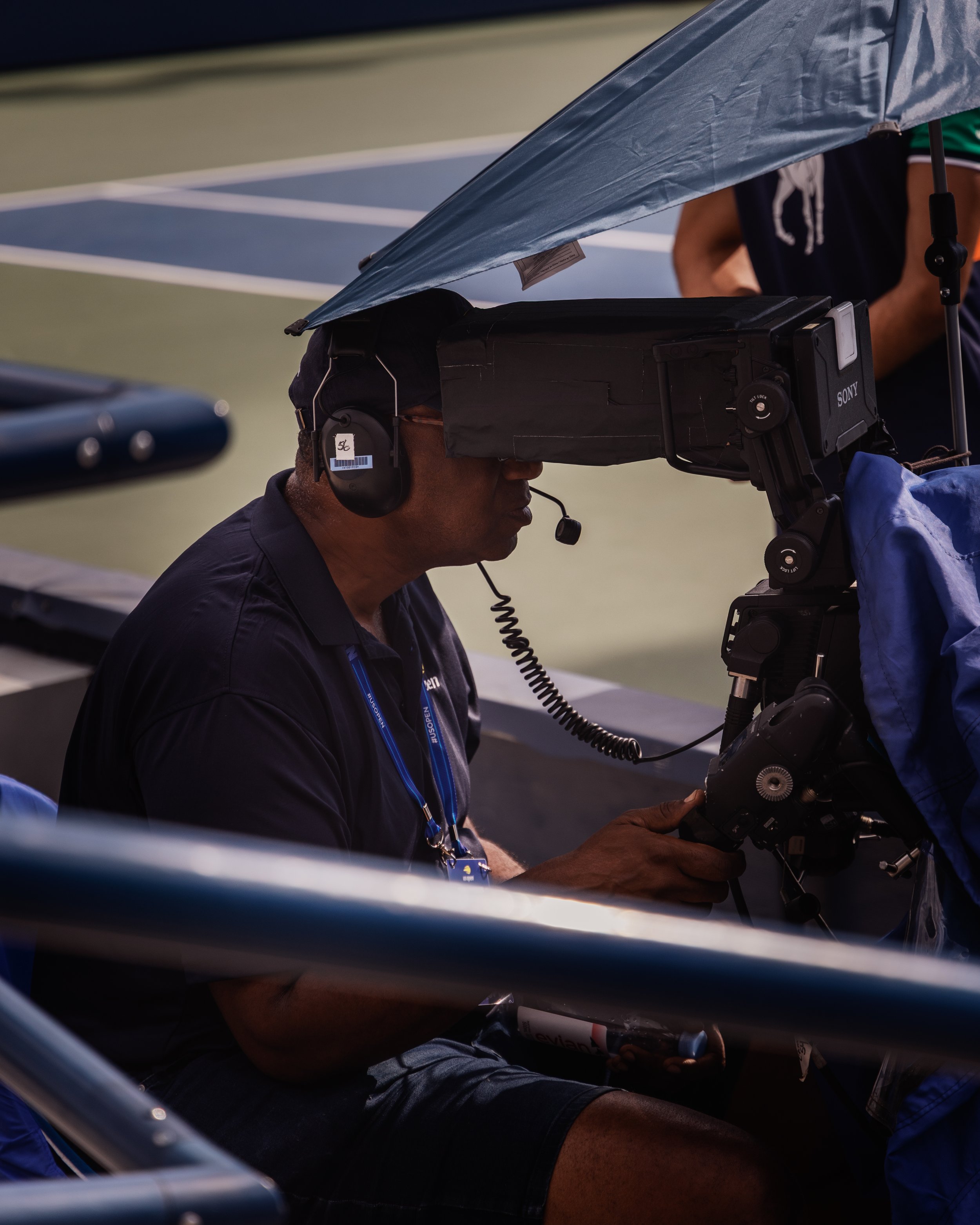 Un homme utilisant une caméra vidéo sophistiquée dans un stade ou une arène, probablement pour filmer un événement sportif ou un film. Il porte un casque avec un identité tag et se trouve à côté d'une voiture ou d'une autre structure.