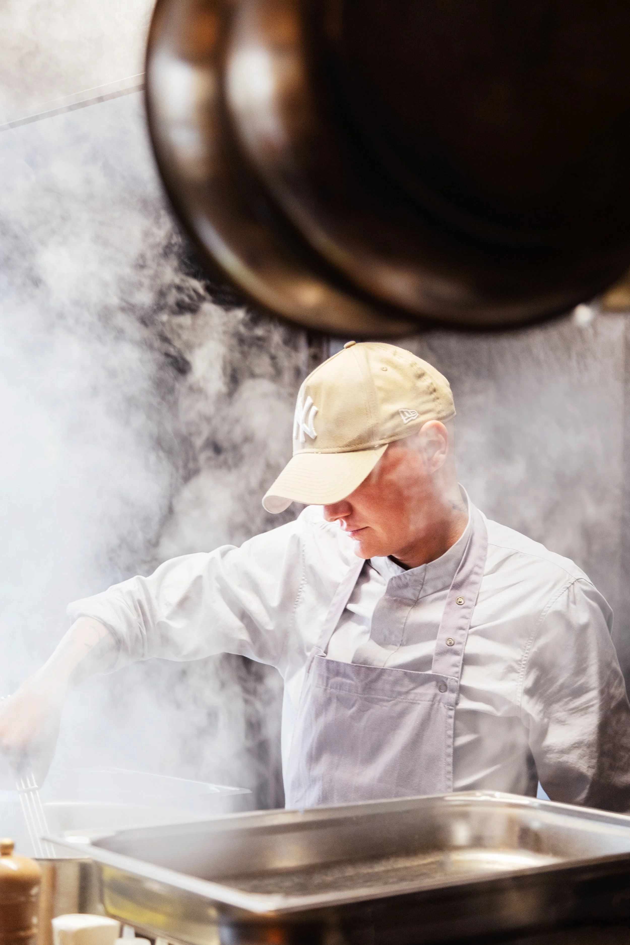 Un chef en cuisine portant une tenue blanche et une casquette beige, en train de cuisiner dans une cuisine avec de la vapeur ou de la fumée.