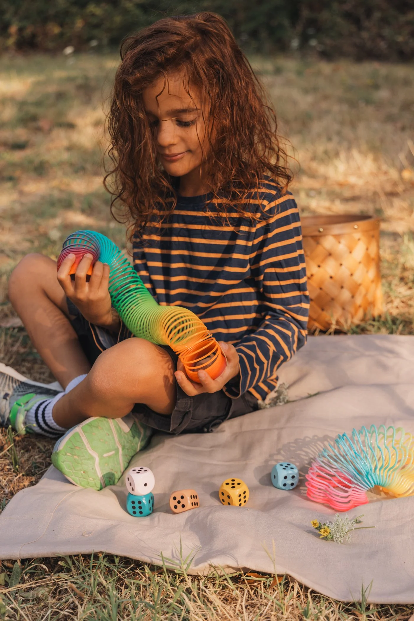 Une jeune fille rousse assise sur un tapis en plein air, jouant avec un jeu de construction en spirale colorée. Elle porte un sweat à rayures orange et bleu, des baskets vert clair, et des chaussettes à rayures. Autour d'elle, il y a des dés colorés et une petite orchidée. En arrière-plan, il y a un panier en osier et la nature.