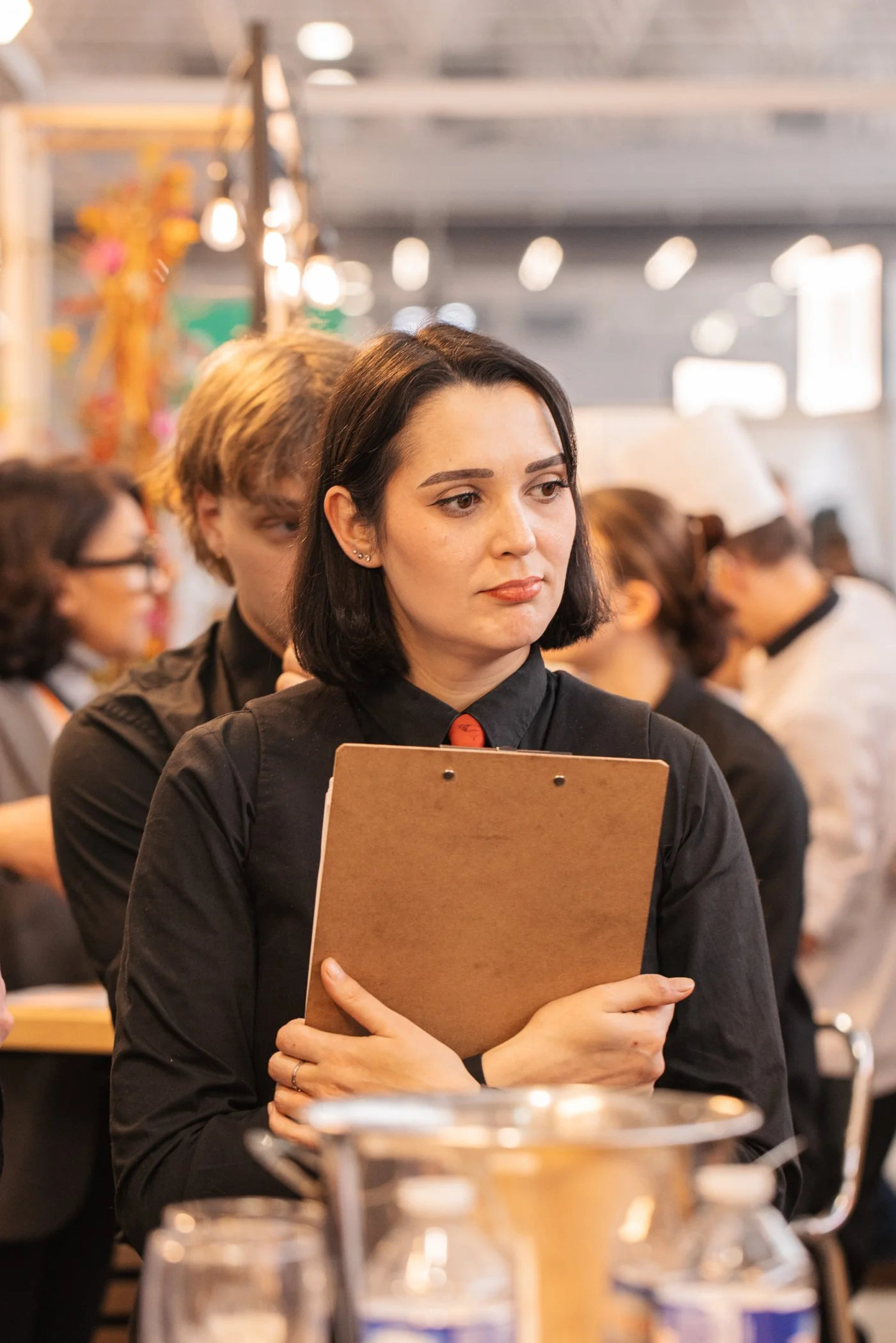 Femme avec cheveux noirs tenant un carnet en bois, portant un uniforme noir avec une bouton rouge, dans un environnement animé avec des collègues en arrière-plan.