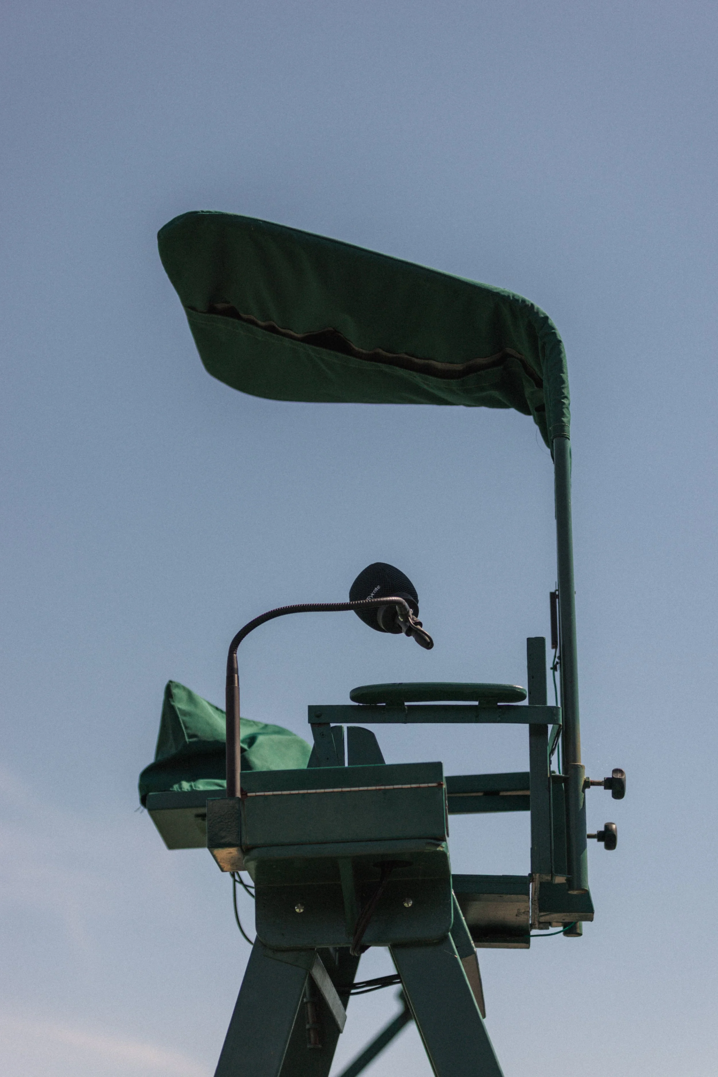 Un drapeau vert flottant dans le vent avec un ciel bleu en arrière-plan, placé sur une structure métallique en haut d'une tour ou d'une plateforme, accompagnée d'un microphone et d'une autre structure métallique.