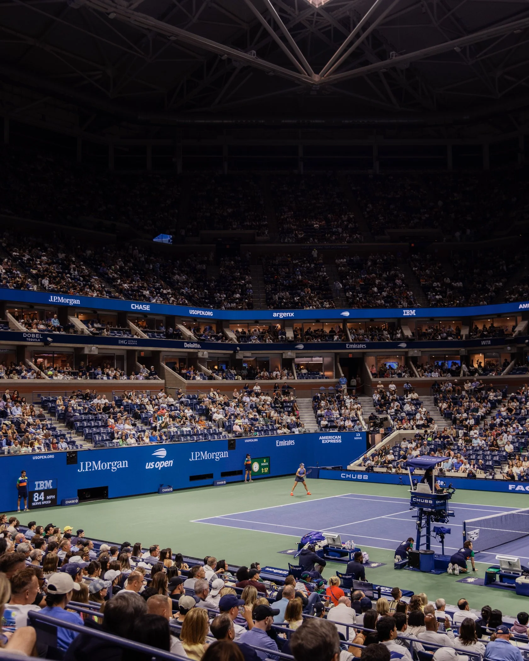 Une scène de match de tennis à l'intérieur d'un stade, avec des spectateurs assis dans les gradins et un court de tennis au centre. Des écrans publicitaires entourent le court, et il y a des caméras de tournage sur le côté.