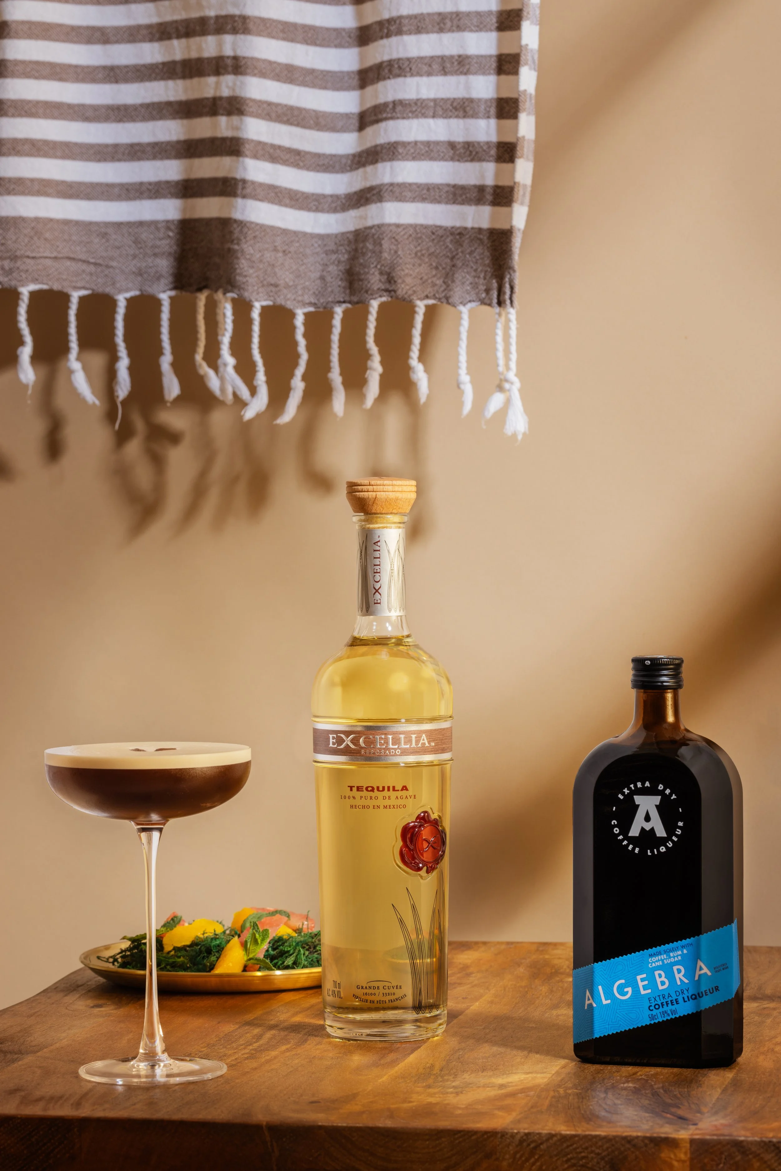 Une bouteille de tequila, une bouteille de liqueur noire, un verre à cocktail avec une boisson brun clair, une assiette de nourriture colorée et une serviette avec des rayures horizontales krem et blanc suspendue contre un mur beige.