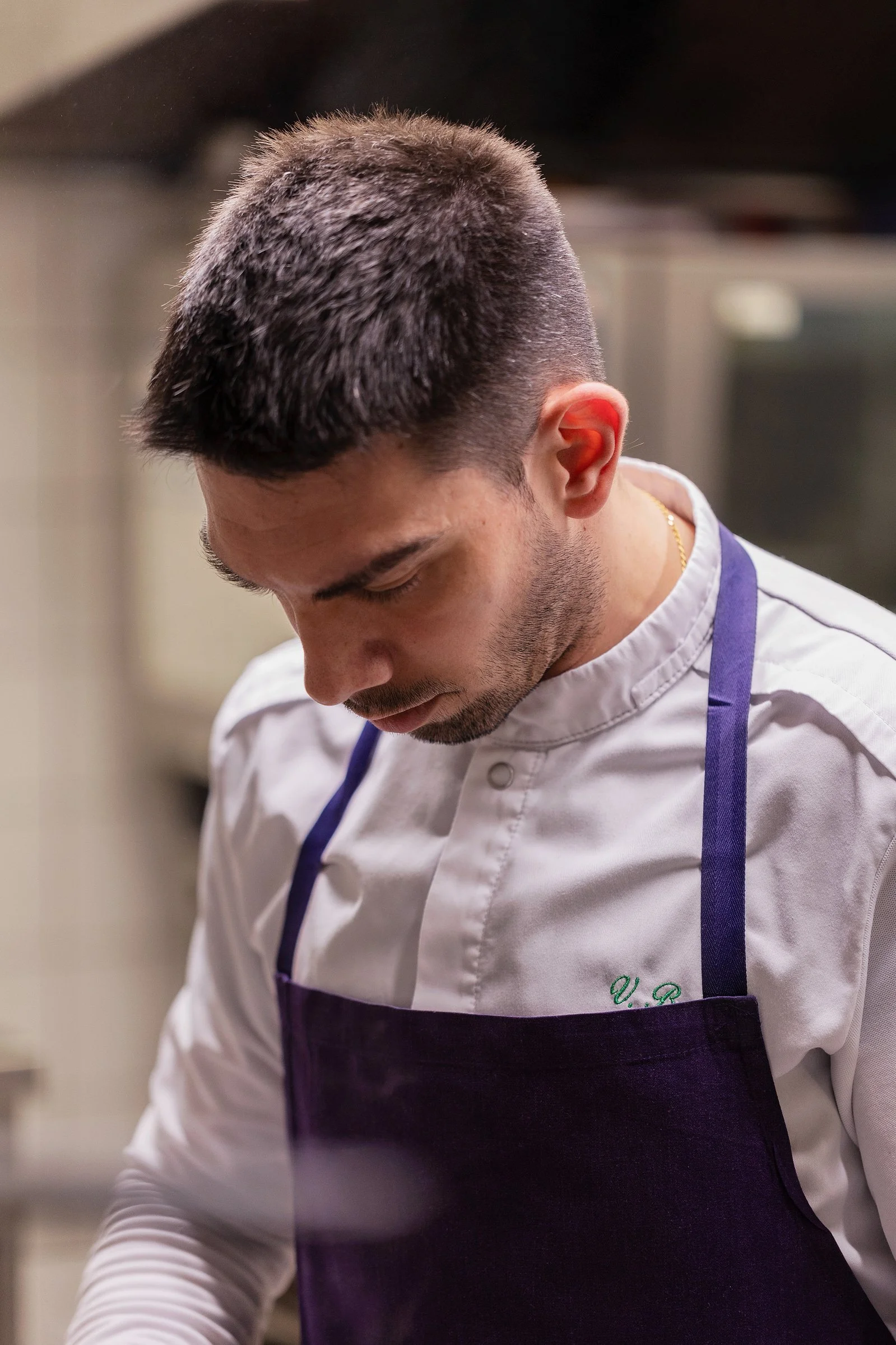 Un homme en uniforme de cuisine, portant un tablier violet, vue de dos, se concentrant sur une tâche. 