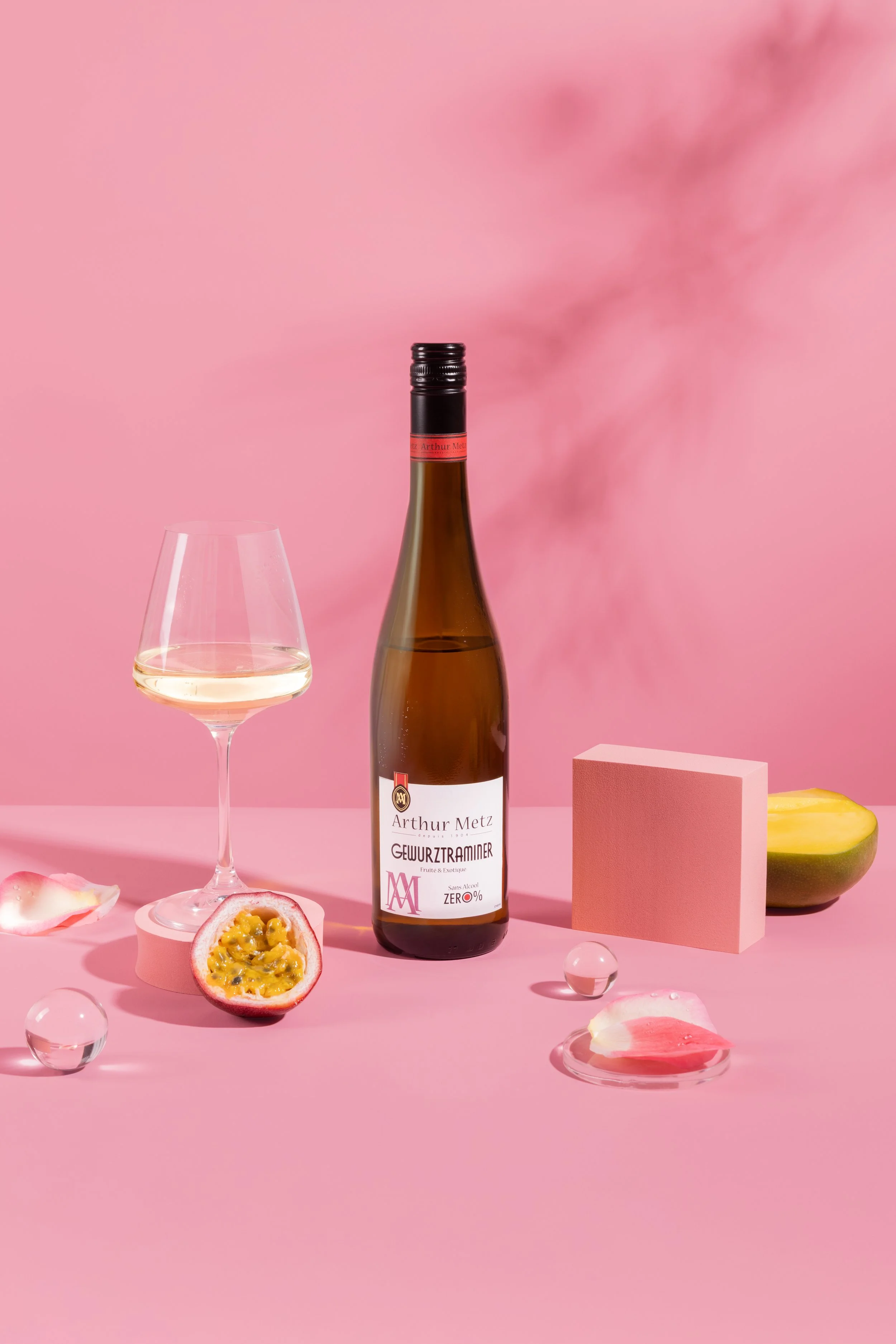 Une bouteille de vin avec un verre de vin blanc, un fruit de passion coupé, des pétales de fleur et des boules en verre, sur un fond rose pastel.