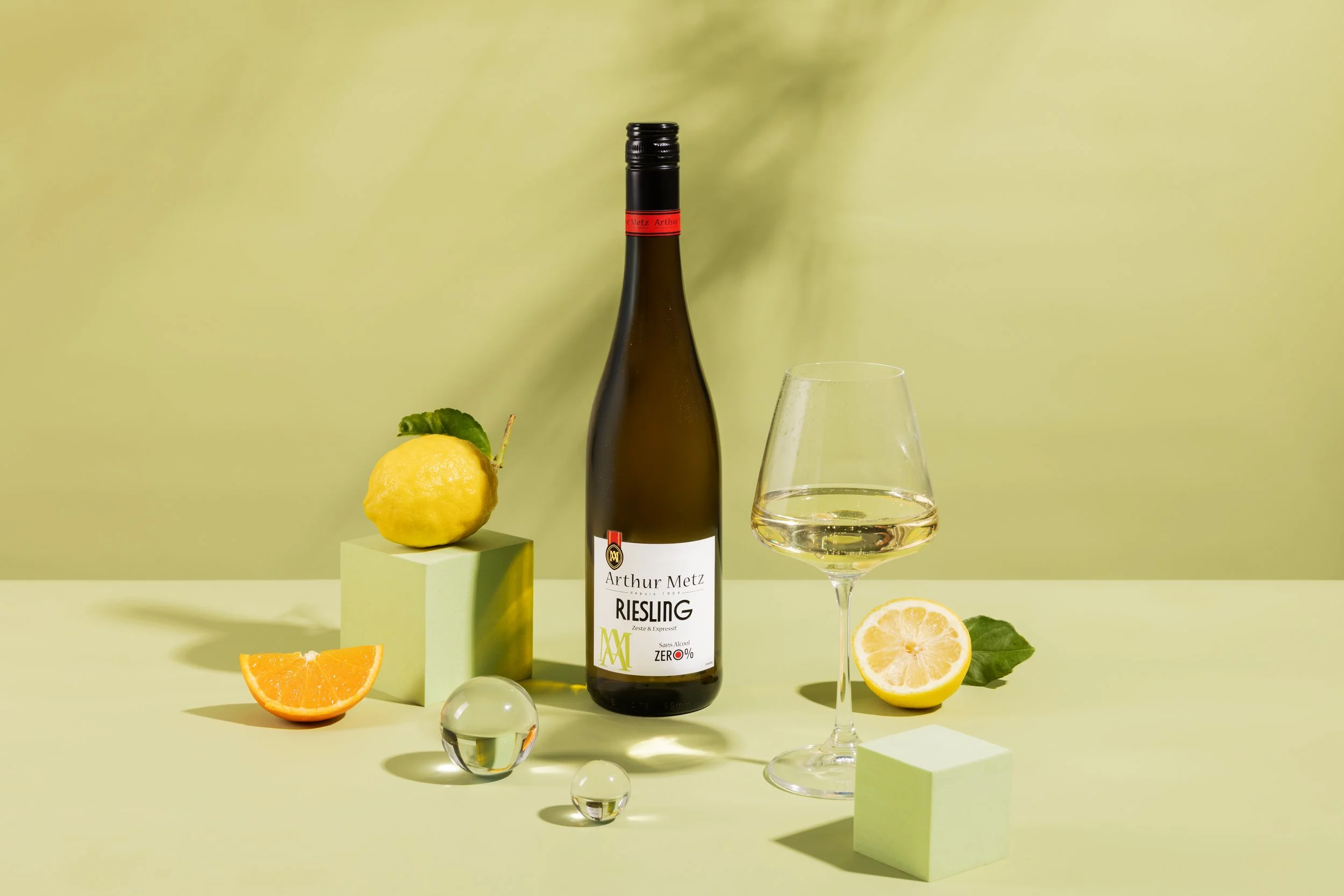 Bouteille de vin Riesling Arthur Metz, un verre de vin blanc, quartiers de citron, d'orange et un citron entier avec une feuille verte, tous disposés sur un fond vert clair avec des blocs de présentation