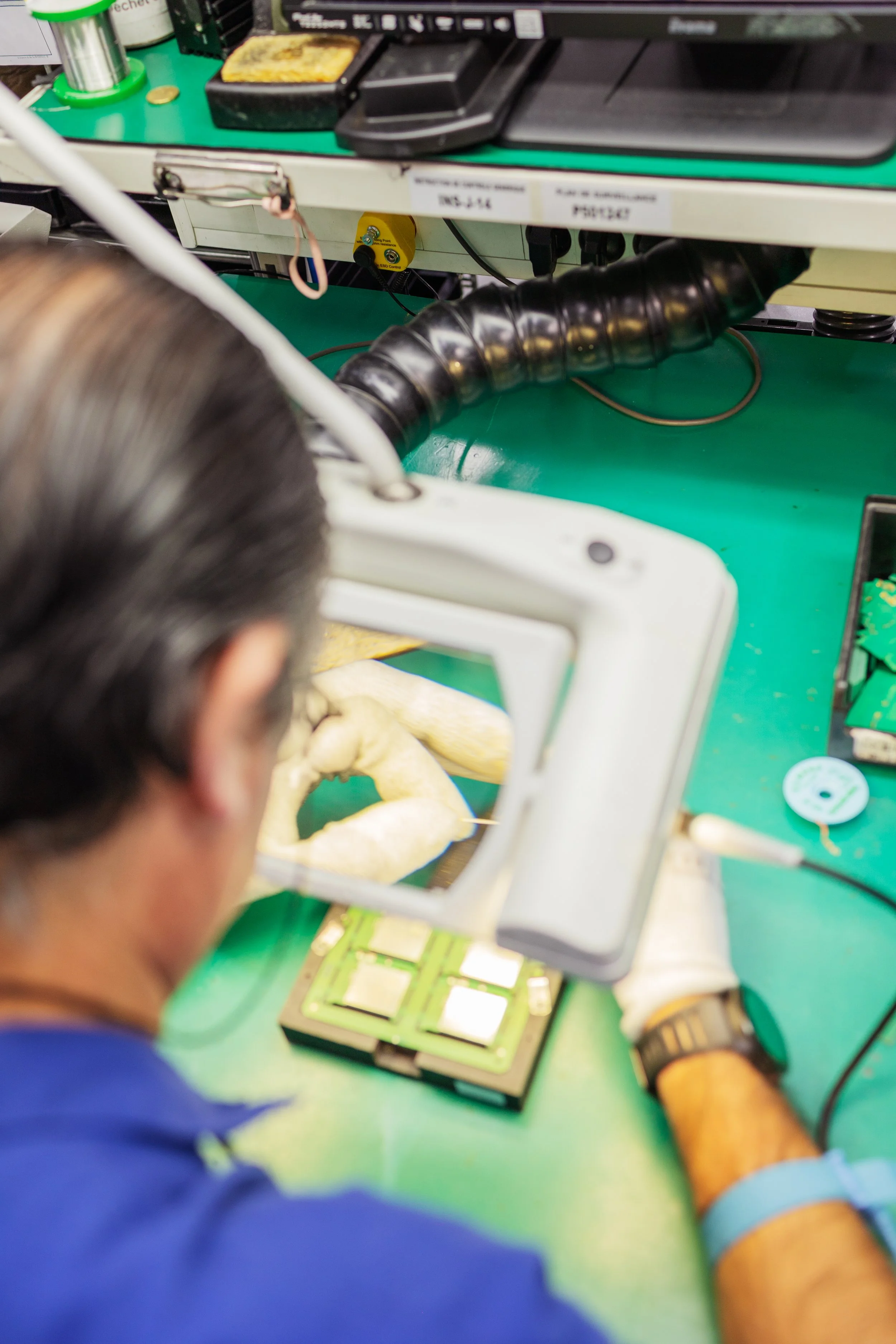 Un technicien en laboratoire utilisant un microscope pour examiner un circuit électronique à un poste de travail, avec divers outils et équipements électroniques autour.