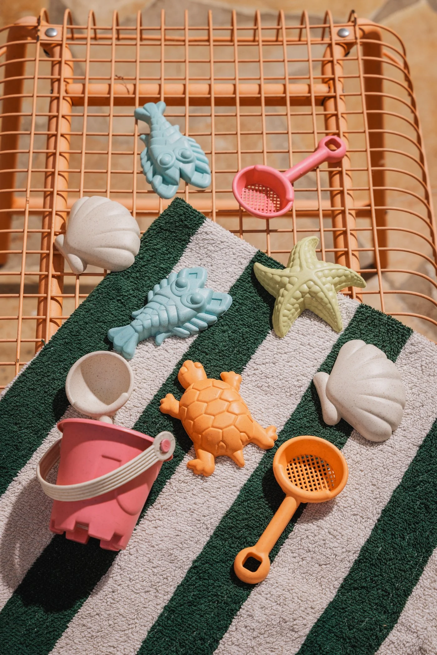 Jeu de plage avec des jouets en plastique, notamment un seau rose, une pelle rose, un filet orange, un crabe blanc, un tortue orange, une étoile de mer jaune, et deux coquillages blancs sur une serviette rayée vert et blanc, avec une grille orange en fond.
