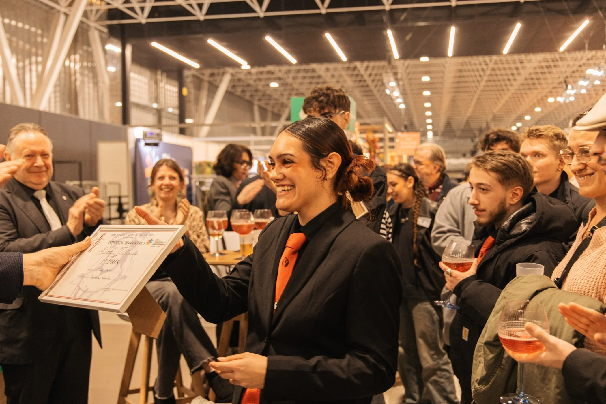 Une cérémonie où une femme reçoit un certificat ou une récompense lors d'un événement, entourée de plusieurs personnes souriantes boitant des verres de vin ou de champagne dans un espace intérieur au plafond éclairé.