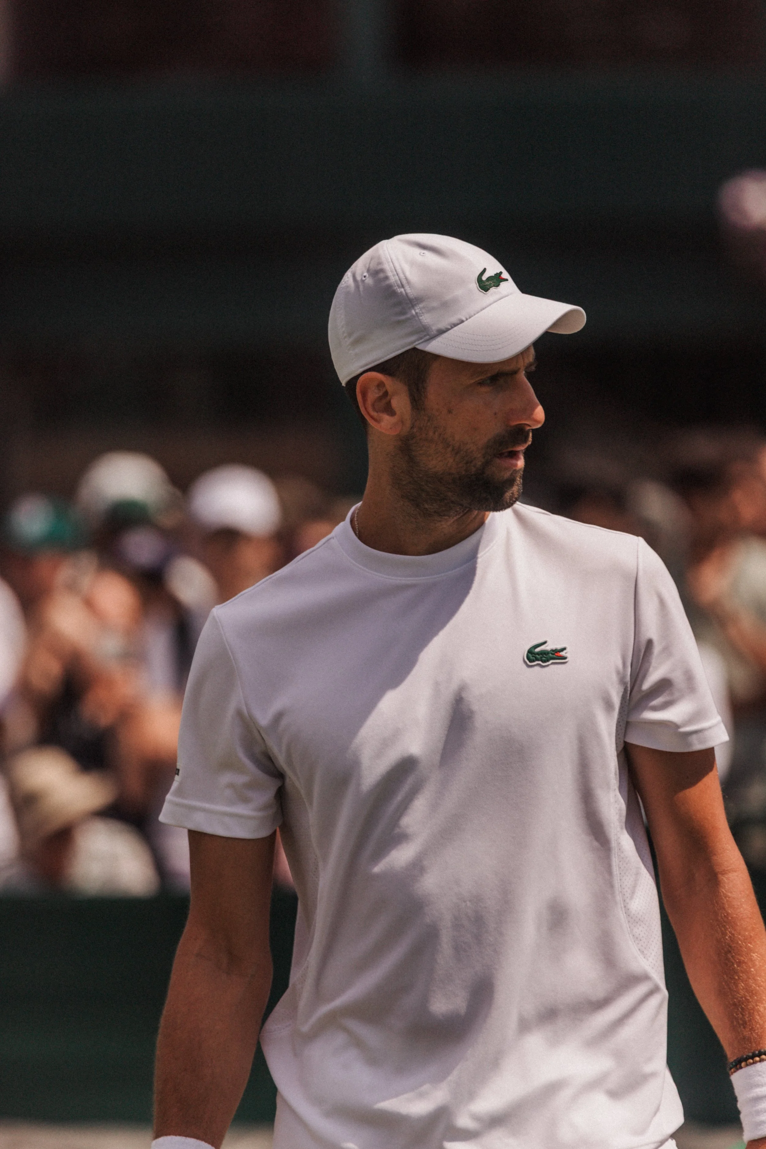 Un homme portant un t-shirt et une casquette blanche avec un logo crocodile vert. Il est en extérieur, probablement lors d'une activité sportive ou un événement, avec une foule floue en arrière-plan.