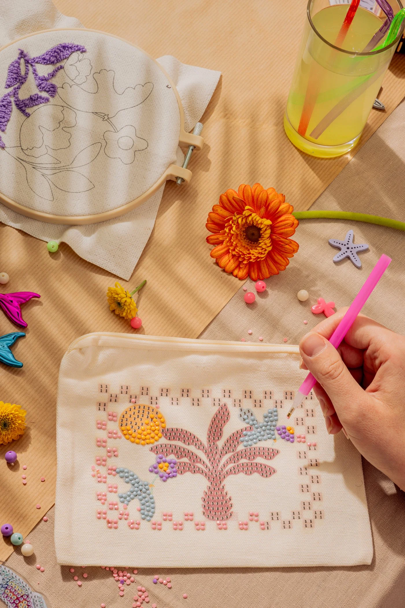 Une personne réalise une broderie diamant représentant un lapin avec des fleurs et des papillons, entourée d'outils, des perles de différentes couleurs et une boisson citronnée avec des pailles.