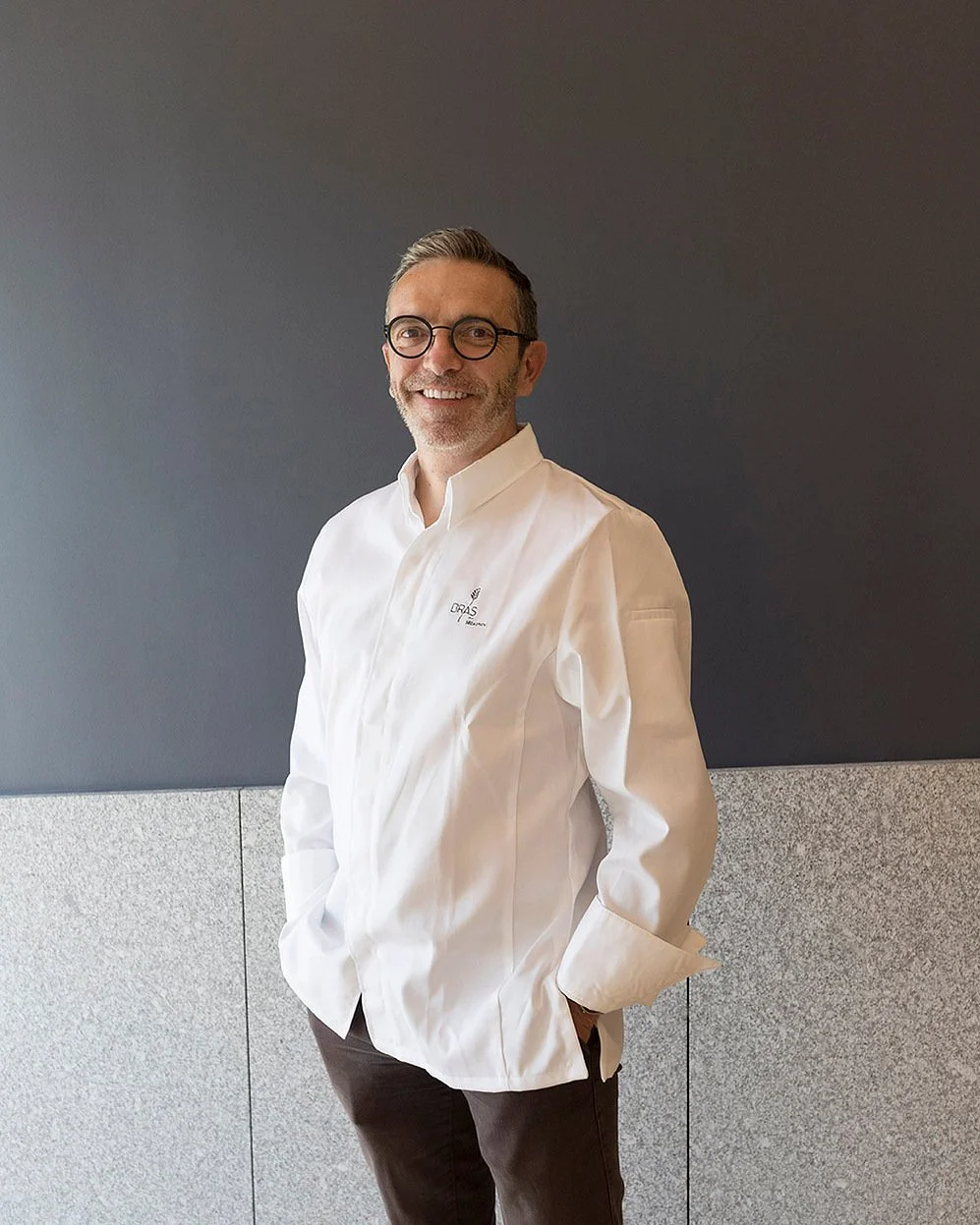 Un homme souriant portant une chemise blanche avec le logo 'ORAS' sur le côté gauche, debout contre un mur gris.
