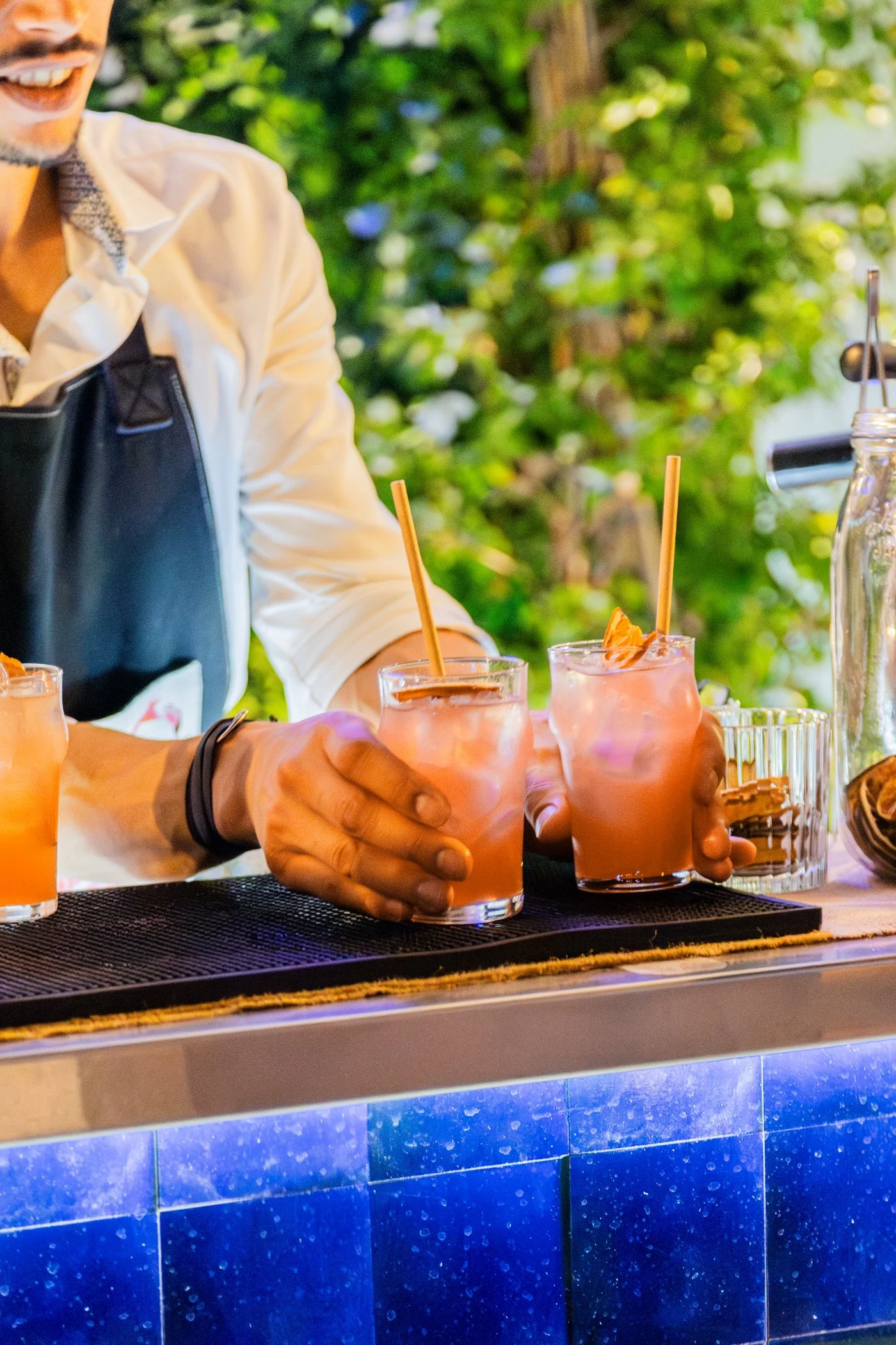 Une personne sert des cocktails roses dans un bar, avec un fond de végétation verdoyante.