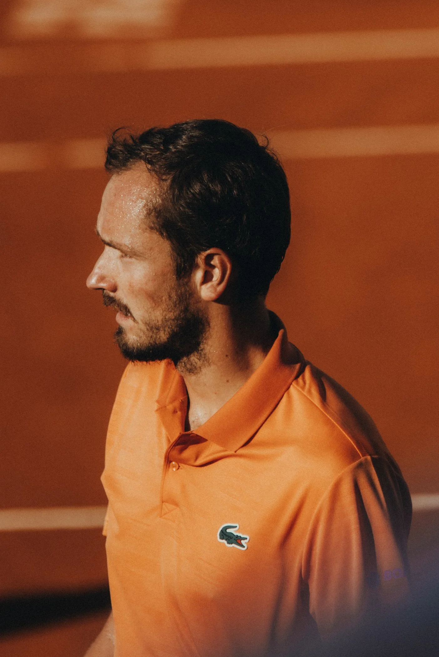 Un homme avec une barbe, portant une chemise orange avec un logo de crocodile Lacoste, vue de profil contre un fond orangé.