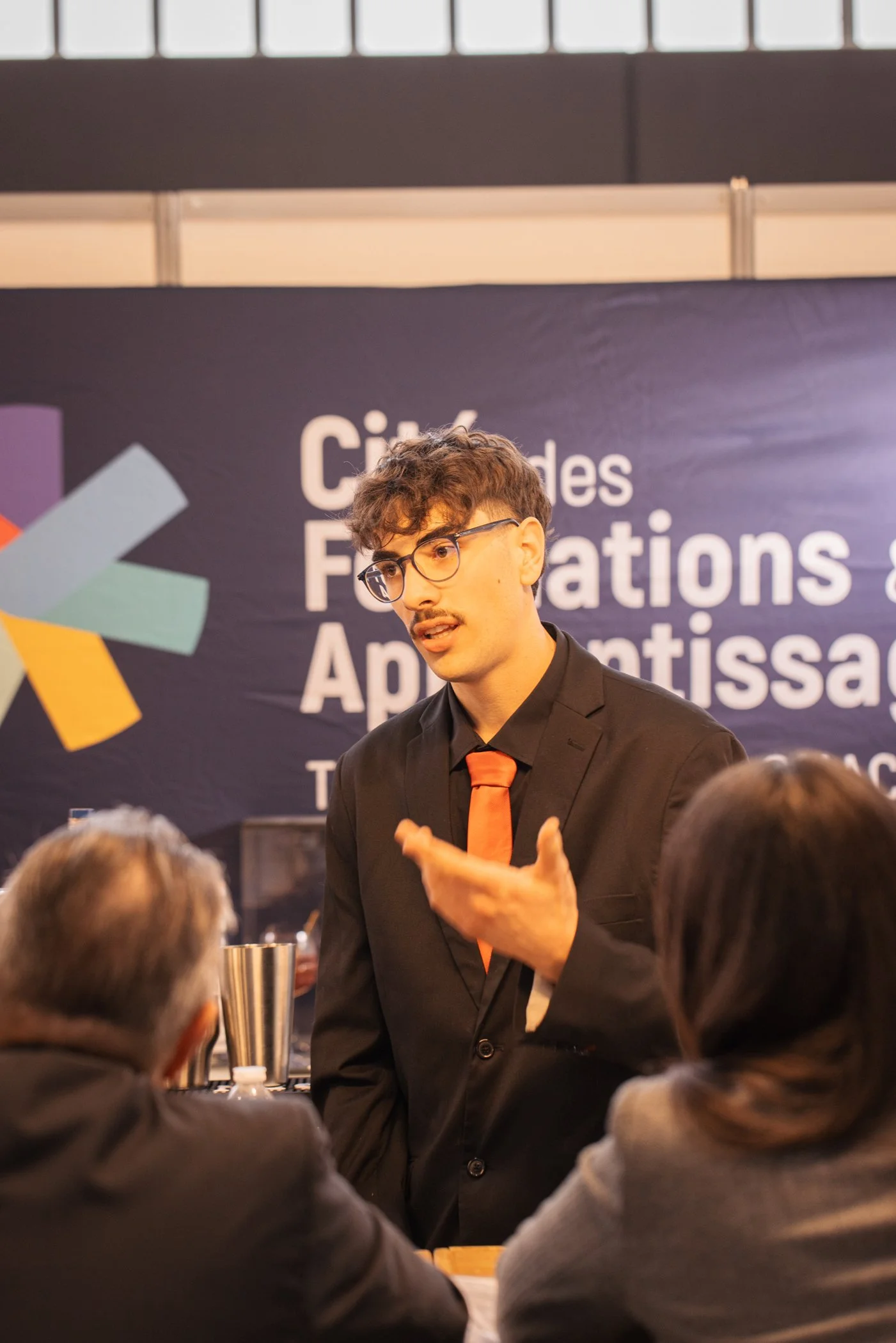 Un homme en costume noir avec une cravate orange parle à un groupe de personnes dans une salle de conférence ou de réunion, avec un panneau en arrière-plan portant le texte en français sur des fondations et applications.