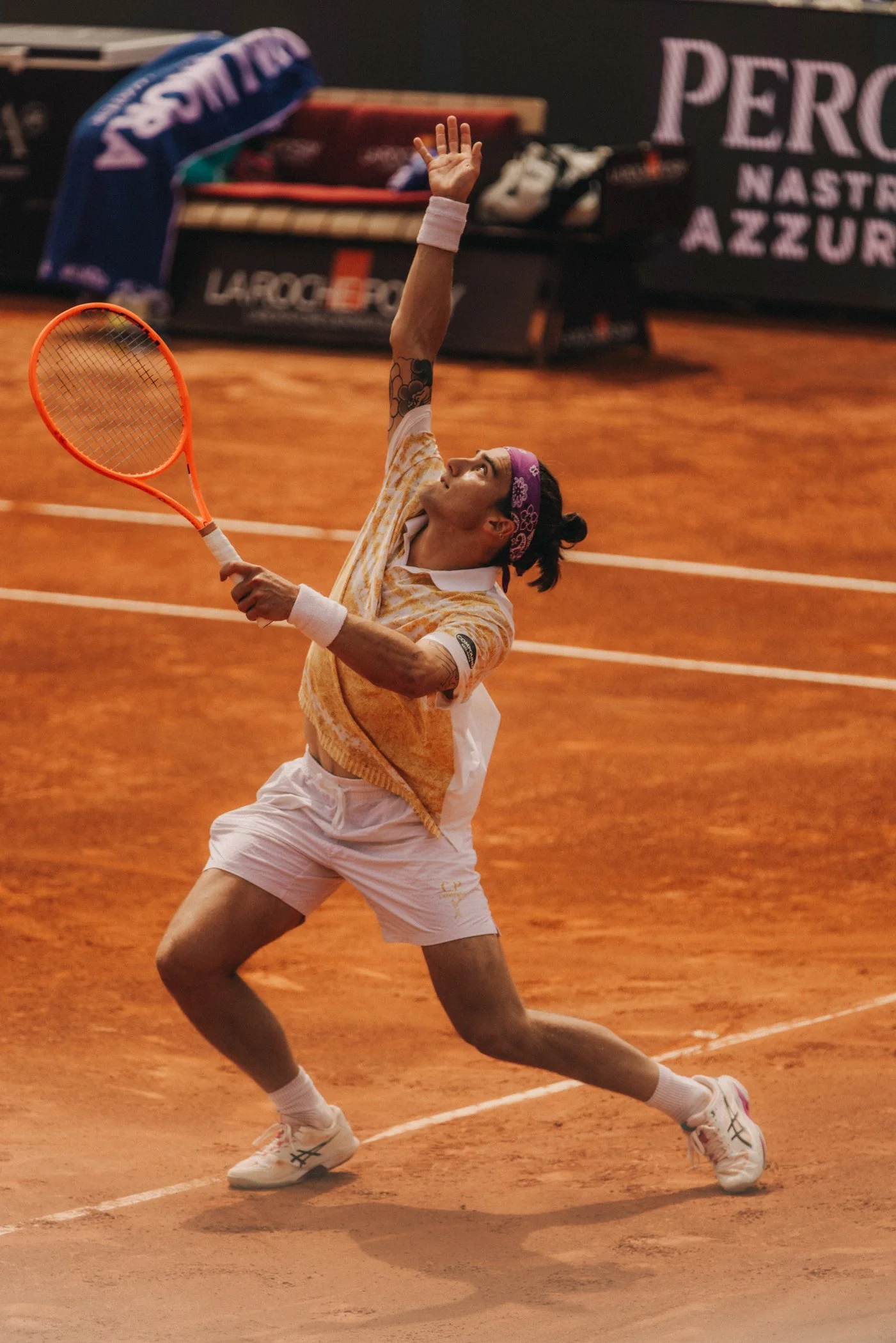 Un joueur de tennis effectue un smash sur un court en terre battue. Il porte un bandeau rose, une chemise orange et blanche, et des chaussures de sport blanches.