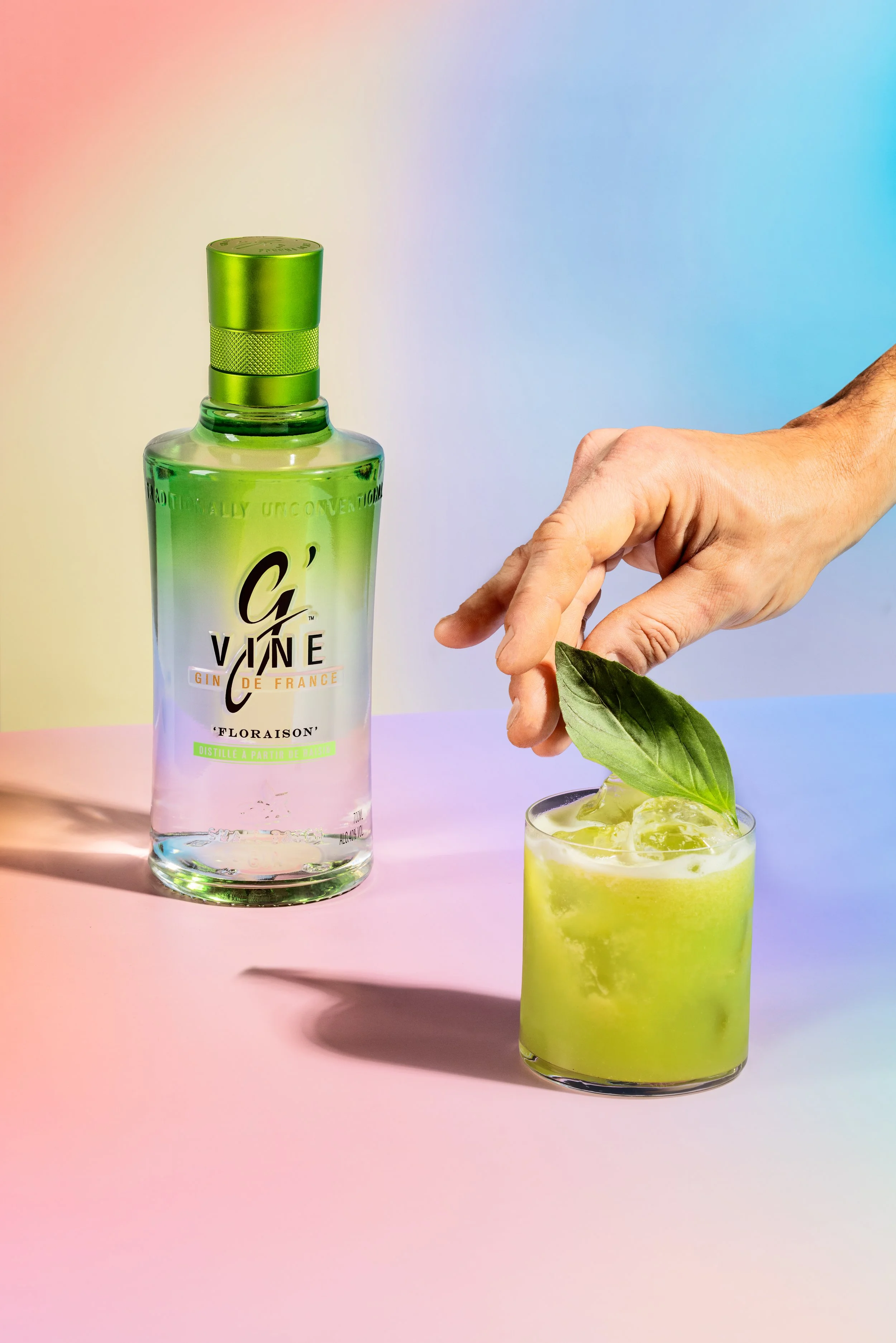 Bouteille de gin vert appelée 'C' VINE', avec un spray vert sur le dessus, à côté d'un verre avec une boisson jaune avec des glaçons, une feuille de basilic et une tranche de citron.