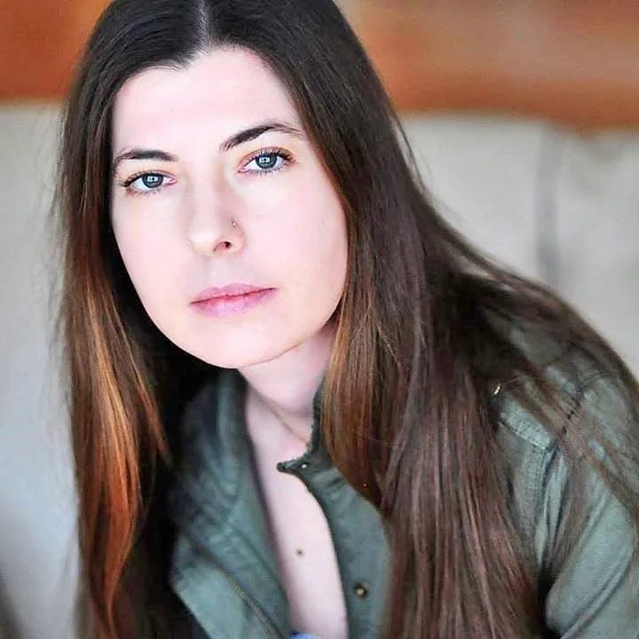 Close-up of a woman with long brown hair and blue eyes, wearing a green jacket and sitting indoors.