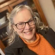 A smiling older woman with glasses and wavy gray hair, wearing an orange sweater and a dark jacket, indoors.