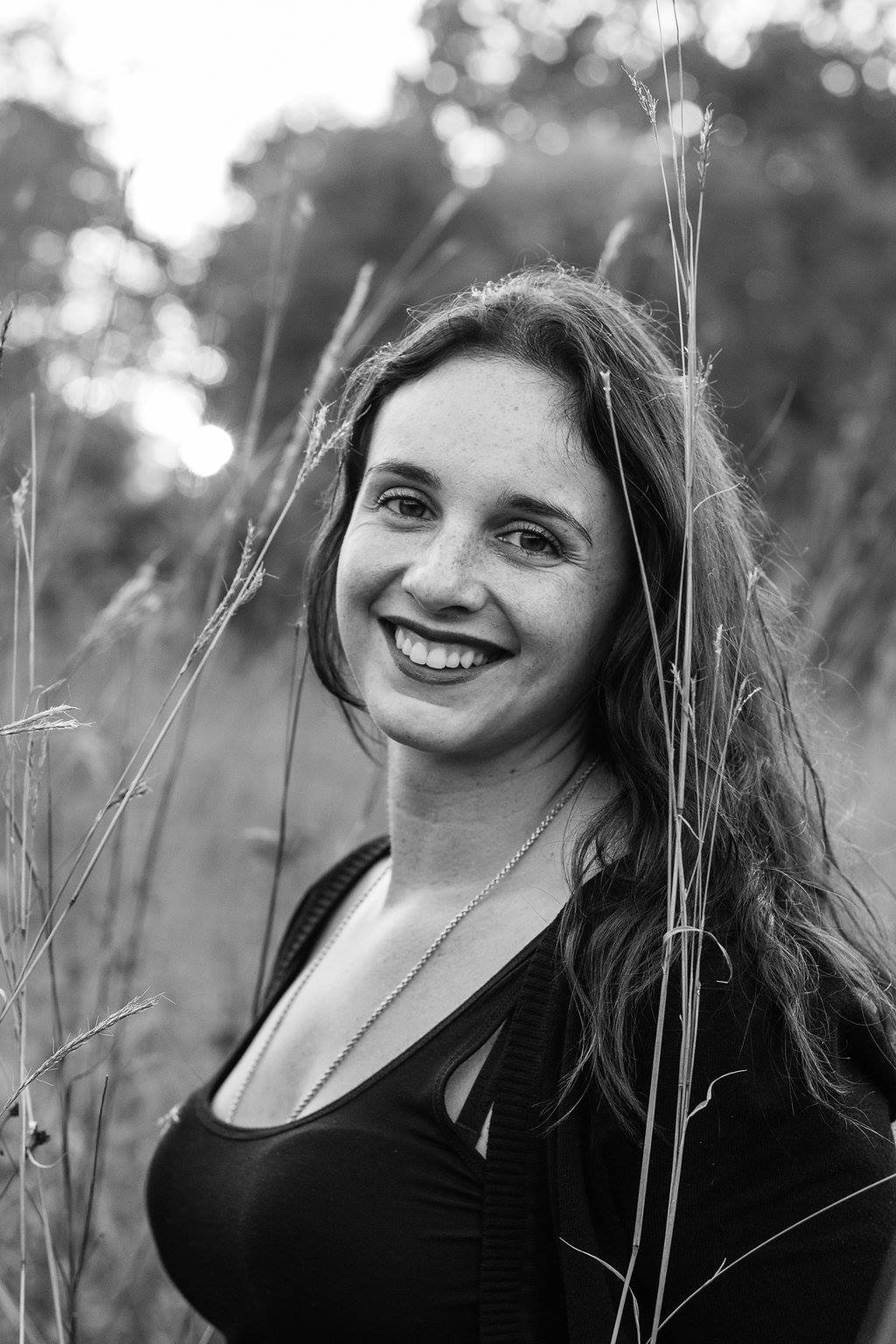 A black-and-white photo of a smiling young woman with wavy hair standing in a field of tall grass.