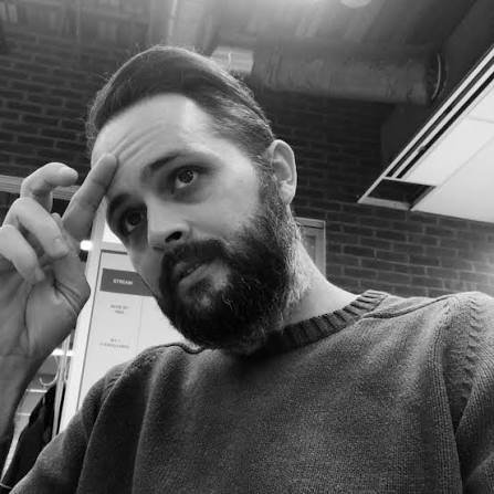 A man with a beard and styled hair sitting indoors, resting his head on his hand with fingers on his forehead, looking thoughtful.