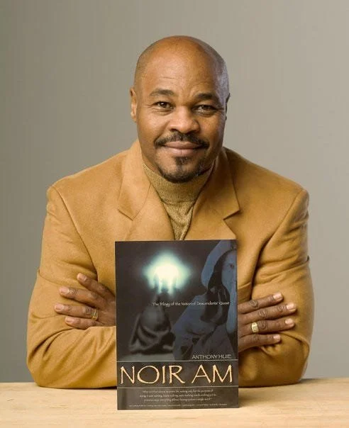 A man with a bald head and a mustache, dressed in a tan blazer and a beige turtleneck, sitting at a table with arms crossed, holding a book titled "Noir Am" in front of him, against a plain light-colored background.