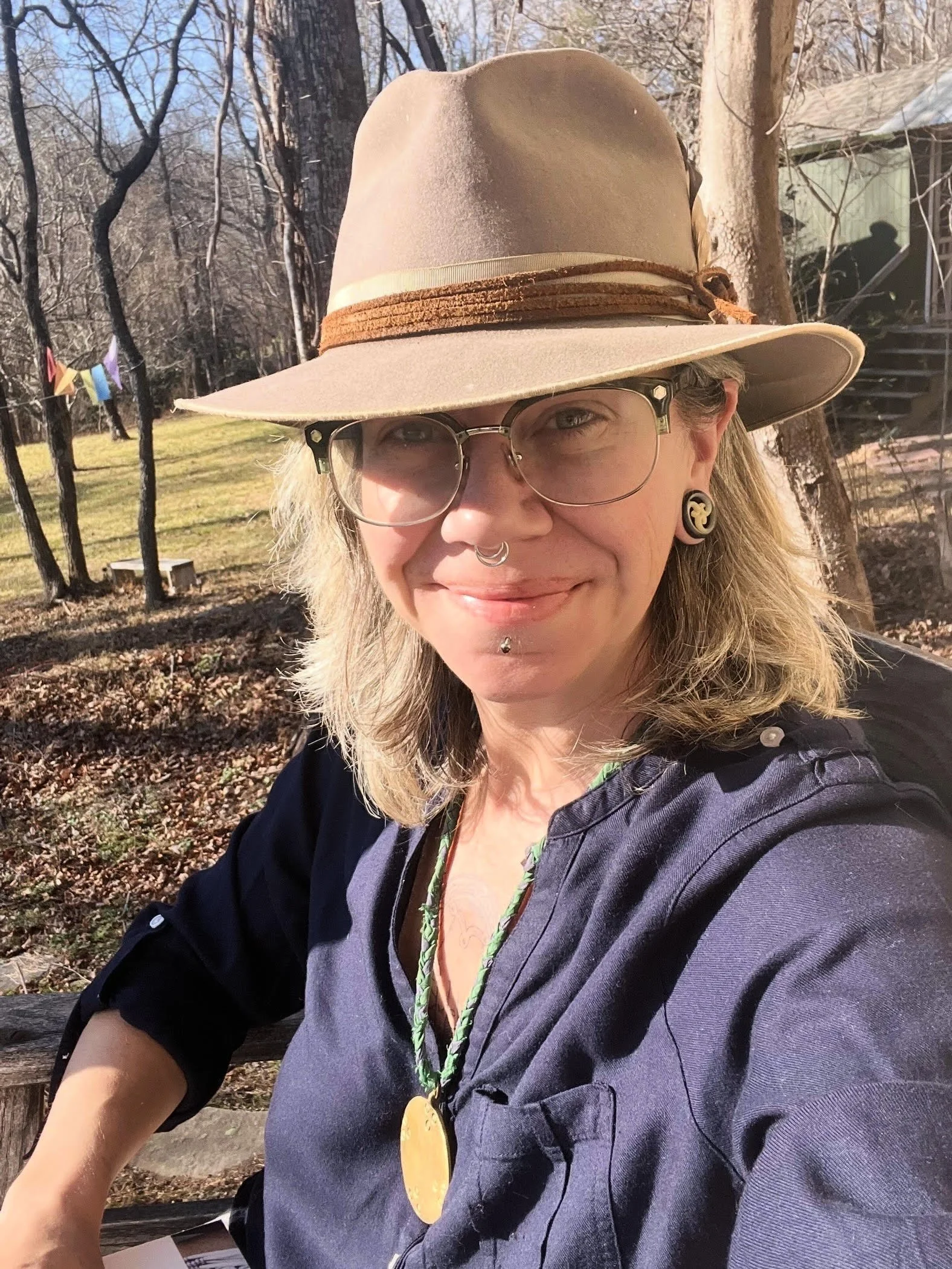 A woman wearing a beige wide-brimmed hat with a brown band, glasses, and earrings, smiling outdoors in a garden with trees and a house in the background.