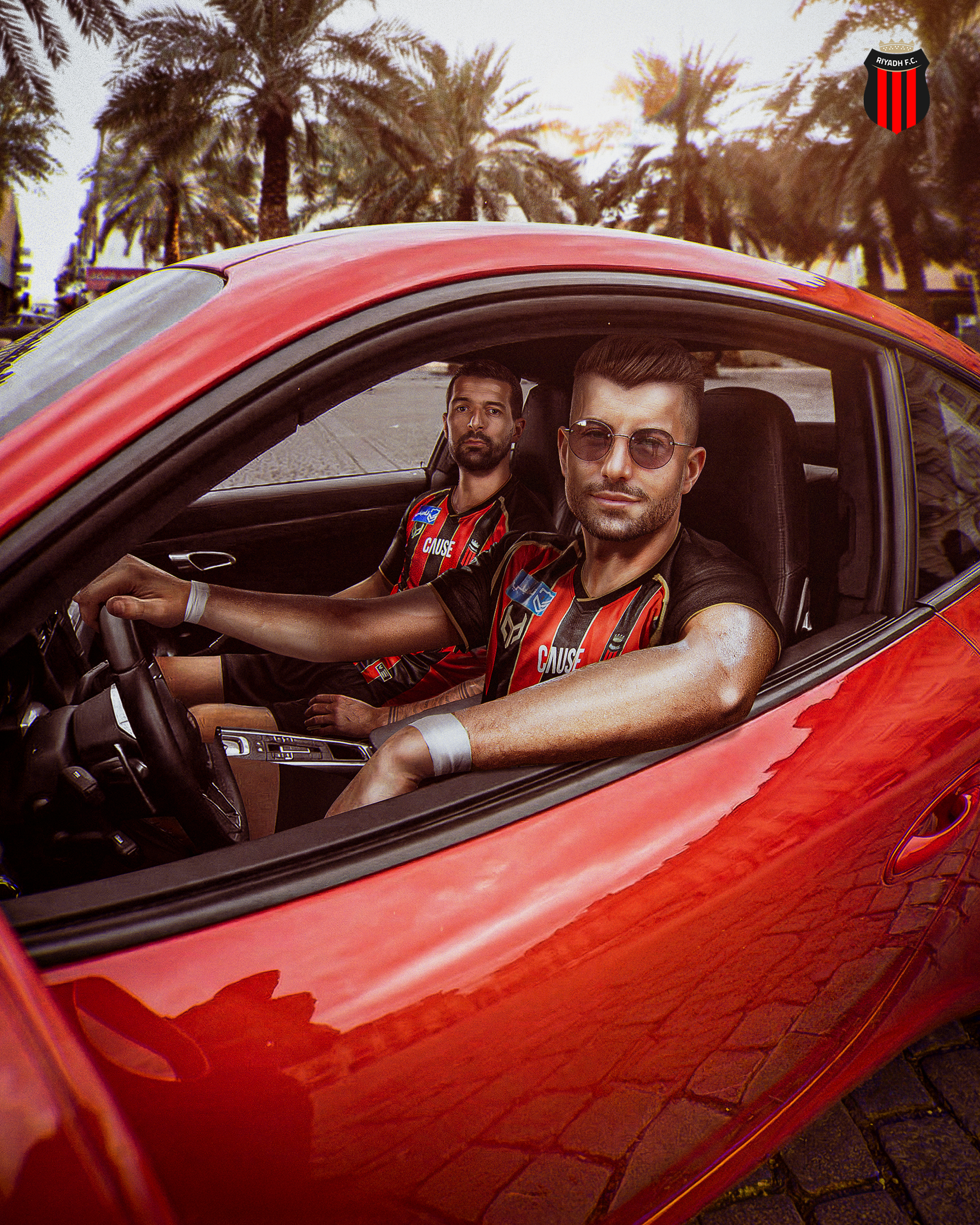 Two men sitting inside a red car with a motorcycle pattern, parked on a cobblestone street lined with palm trees, during sunset, with a black and red Riyadh FC emblem in the top right corner.