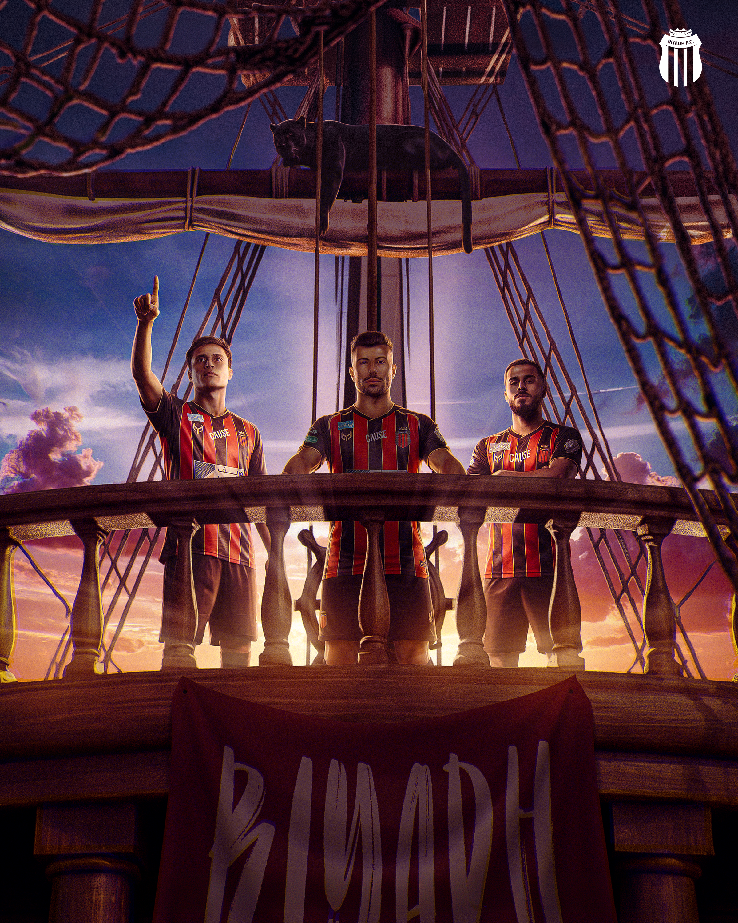 Three men in jerseys with a red, black, and yellow vertical stripe design pose on an outdoor hot air balloon basket at sunset. The basket is made of woven material with a sign hanging in front. The background shows a colorful sky with clouds and the setting sun.