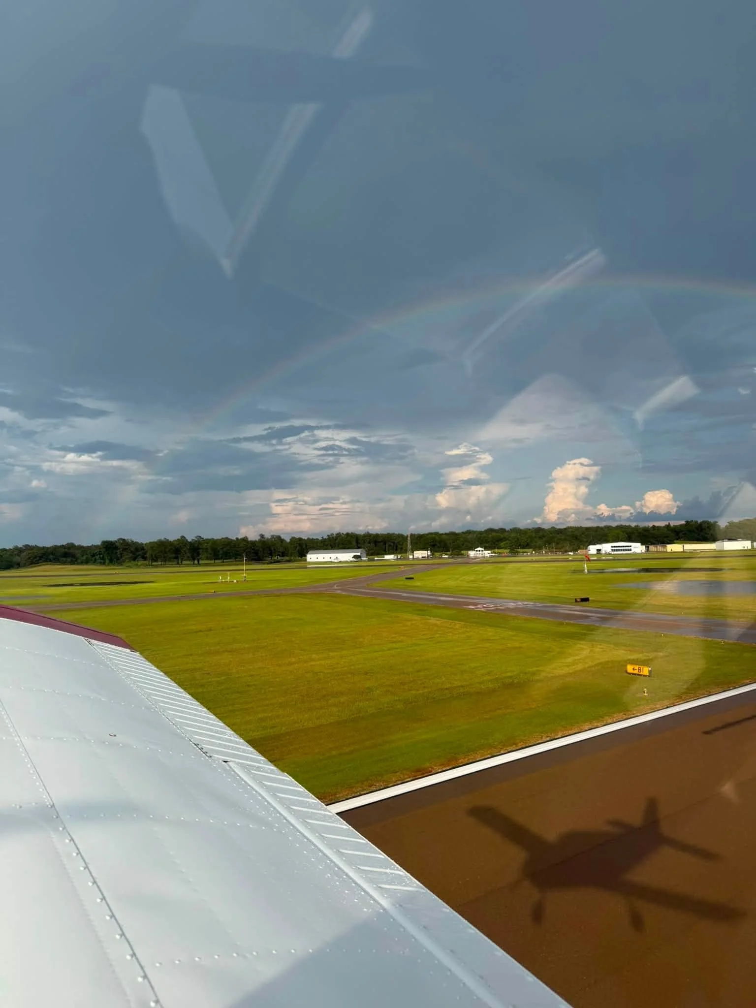 View from an airplane window showing part of the airplane wing with an airport runway below, a grassy field, clouds, and a rainbow in the sky.