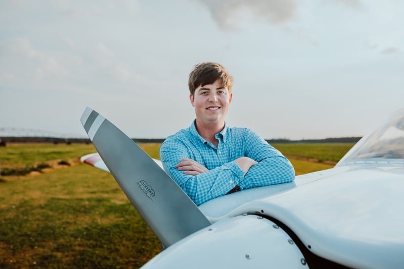 A young man in a blue checkered shirt standing next to an aircraft outdoors, with green fields and a cloudy sky in the background.