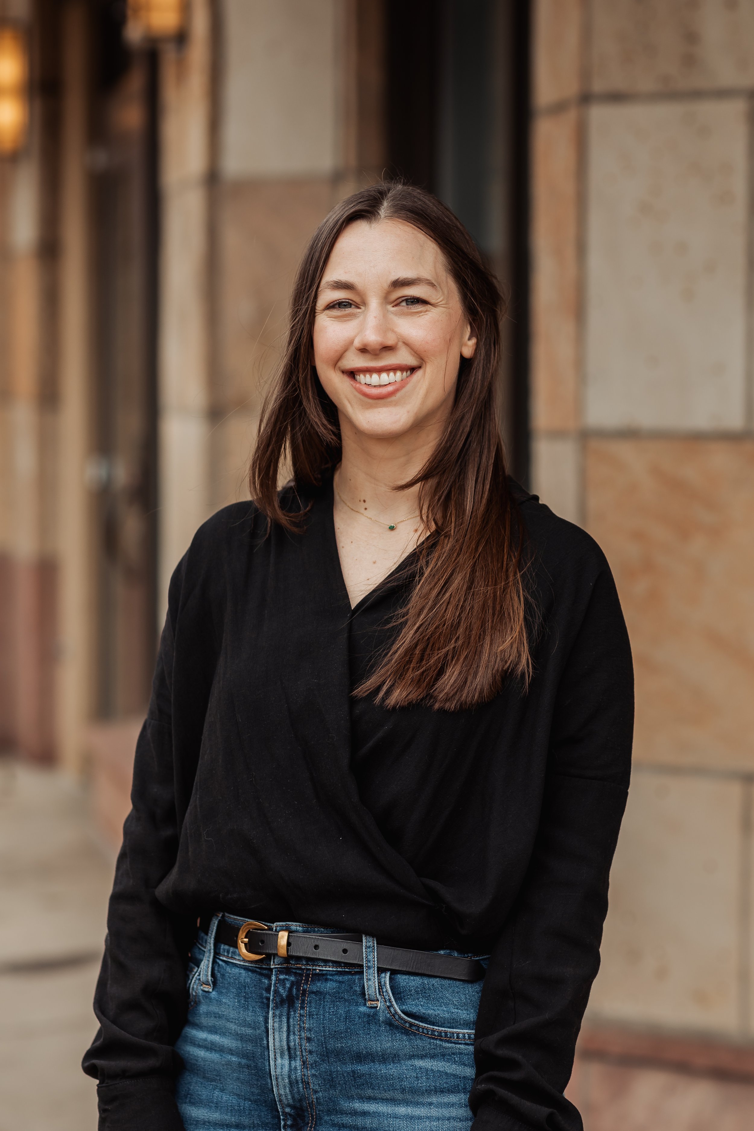 A young woman with long brown hair, Jess Mulligan, smiling and dressed in a black top and blue jeans, standing in front of a brick building in Boulder.