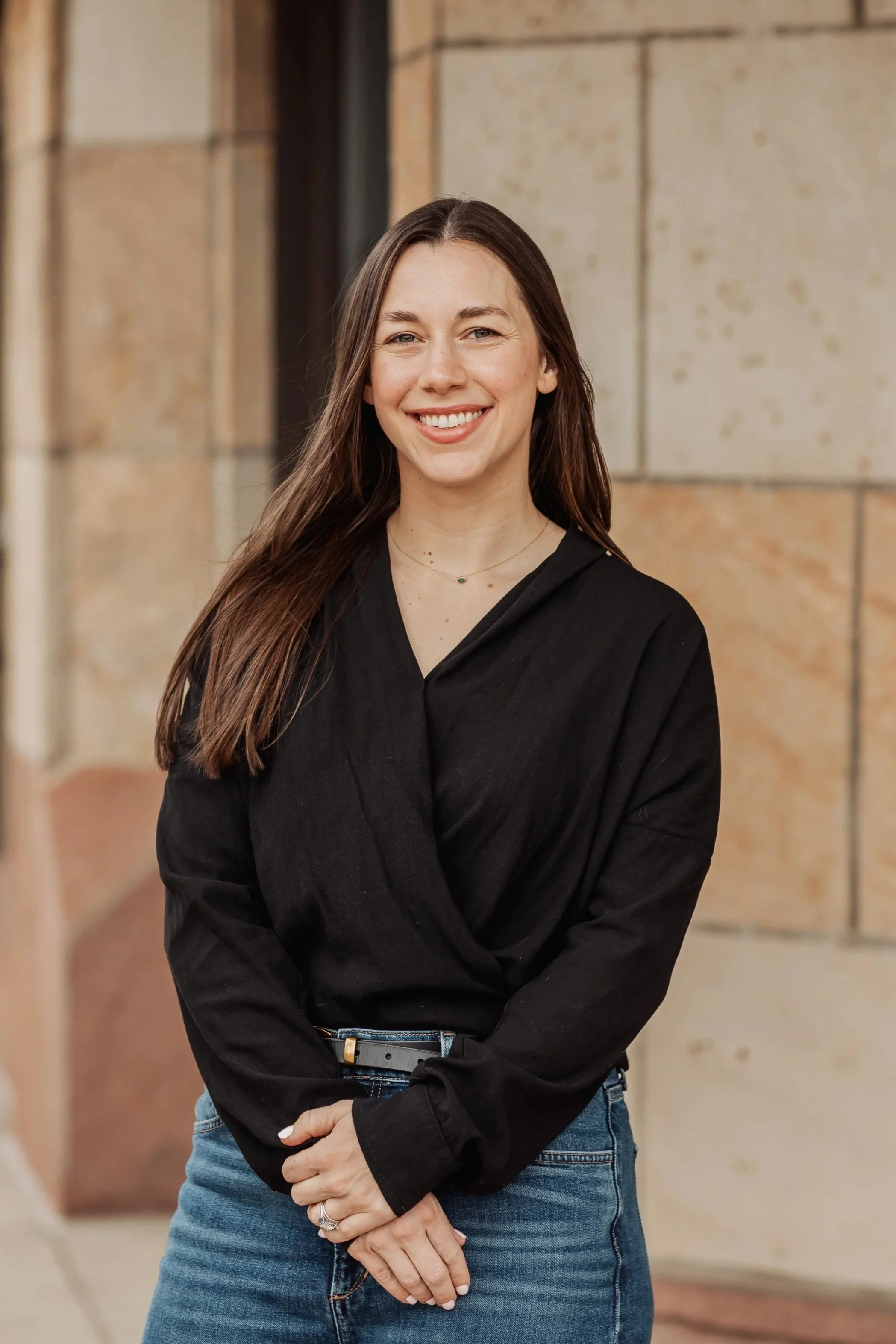 A woman, Jess Mulligan, with long brown hair smiling, wearing a black blouse and blue jeans, standing outside against a beige stone wall.