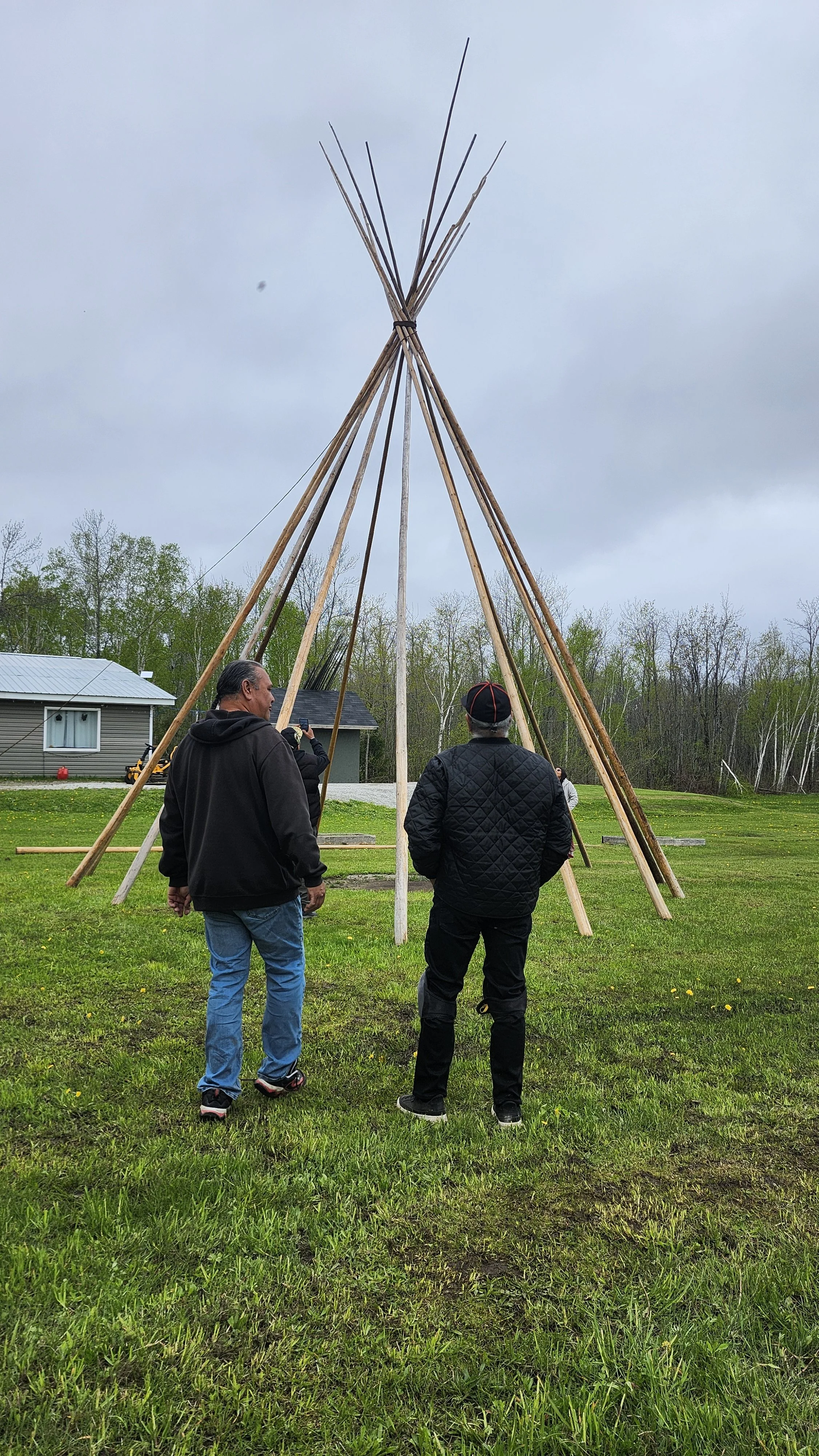 Three men standing on grass looking at a large teepee frame made of wooden poles tied together at the top, with trees and small buildings in the background under a cloudy sky.