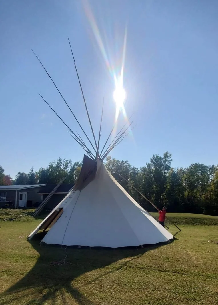 A large teepee-style tent set up outdoors on a grassy area with a person holding one of the supporting poles, bright sun in the sky, trees in the background.